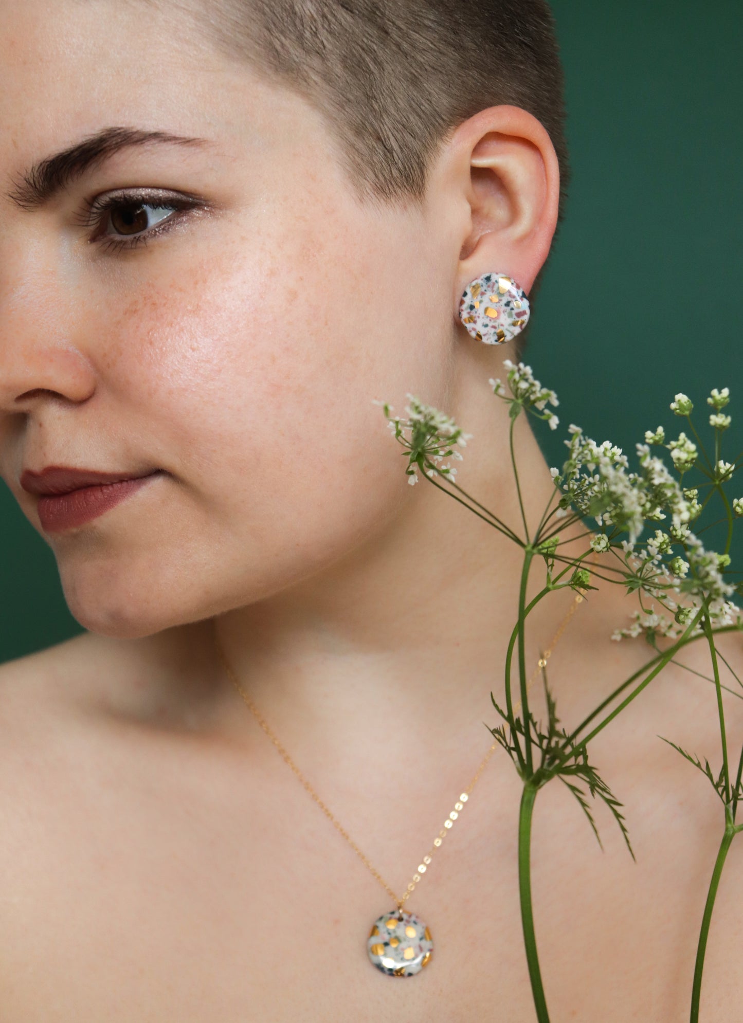 A close-up of a short-haired person wearing the Astilla Recycled Necklace by Rozenthal Jewelry, paired with floral porcelain jewelry, holding white flowers against a green backdrop.