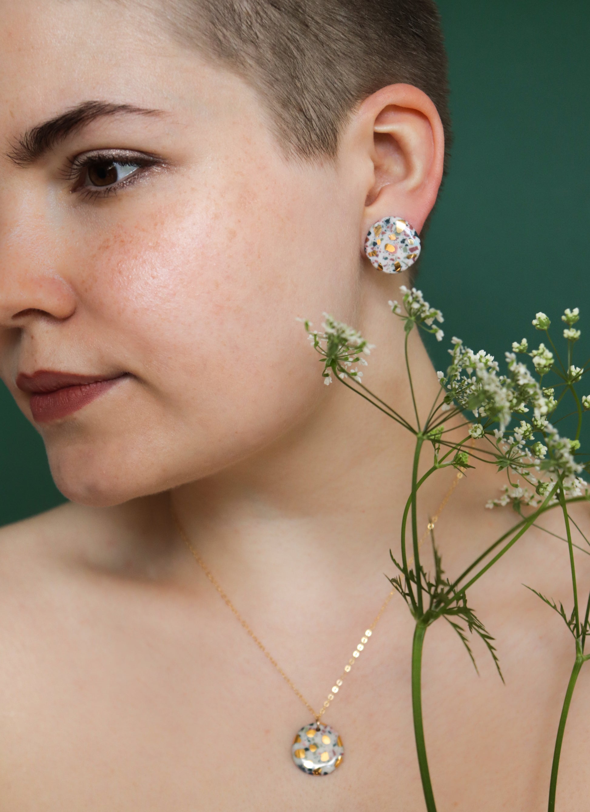 A close-up of a short-haired person wearing the Astilla Recycled Necklace by Rozenthal Jewelry, paired with floral porcelain jewelry, holding white flowers against a green backdrop.