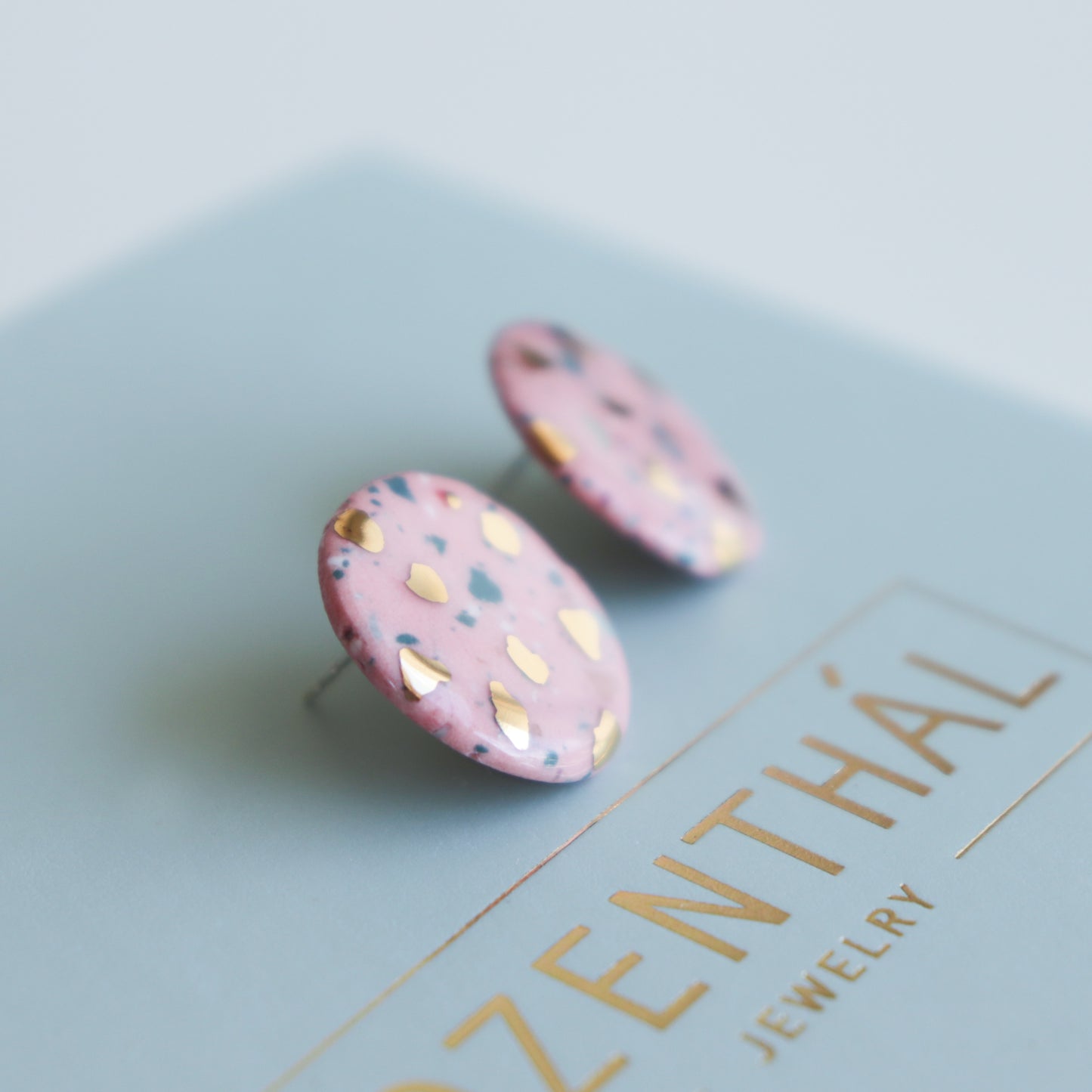 Handmade round pink porcelain earrings with gold and black speckles, the Astilla Recycled Studs / S by Rozenthal Jewelry, displayed on a light blue card.