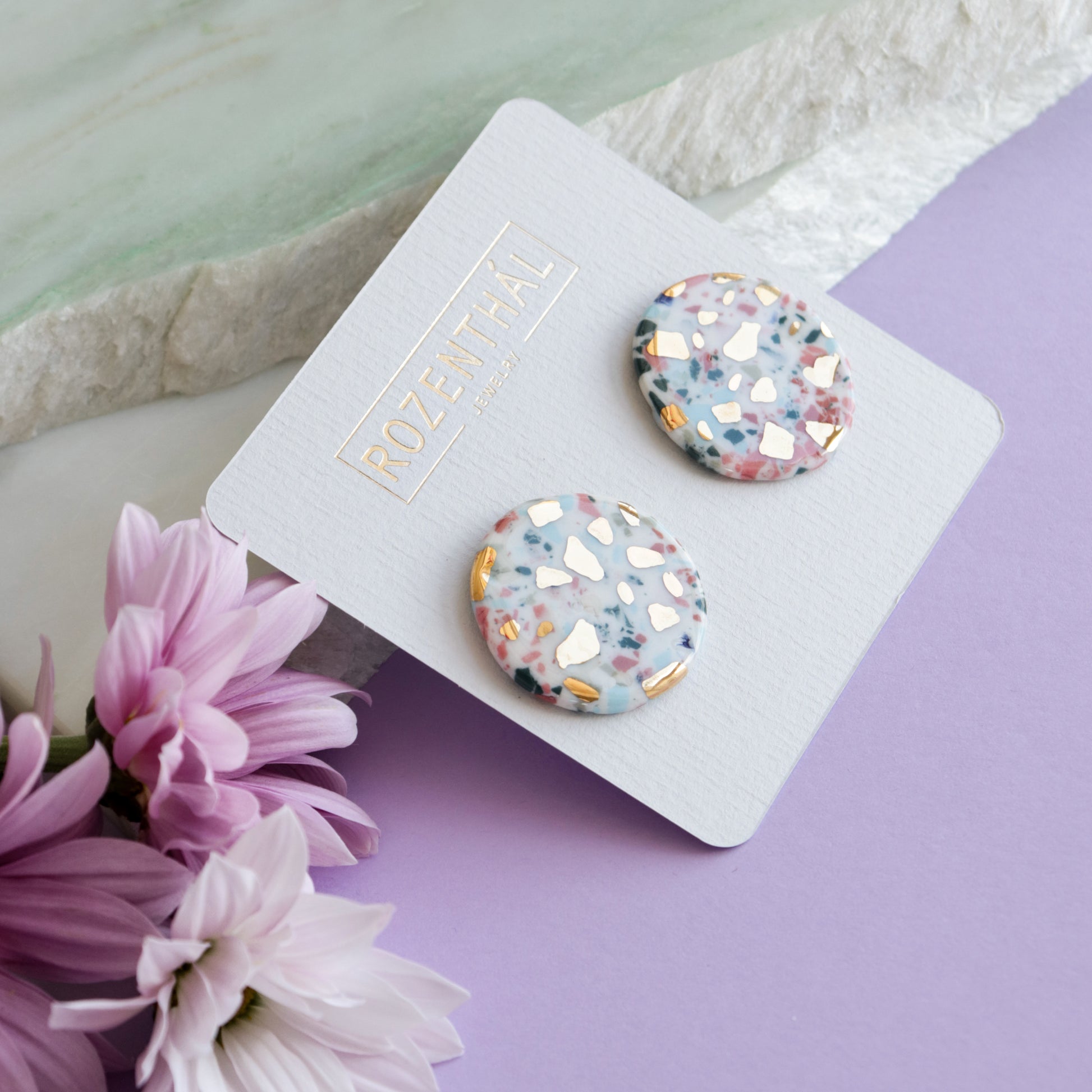 Astilla Recycled Studs / M by Rozenthal Jewelry: handmade round earrings with a pastel terrazzo pattern, shown on a branded card next to pink and white flowers against a lavender-white marble backdrop.