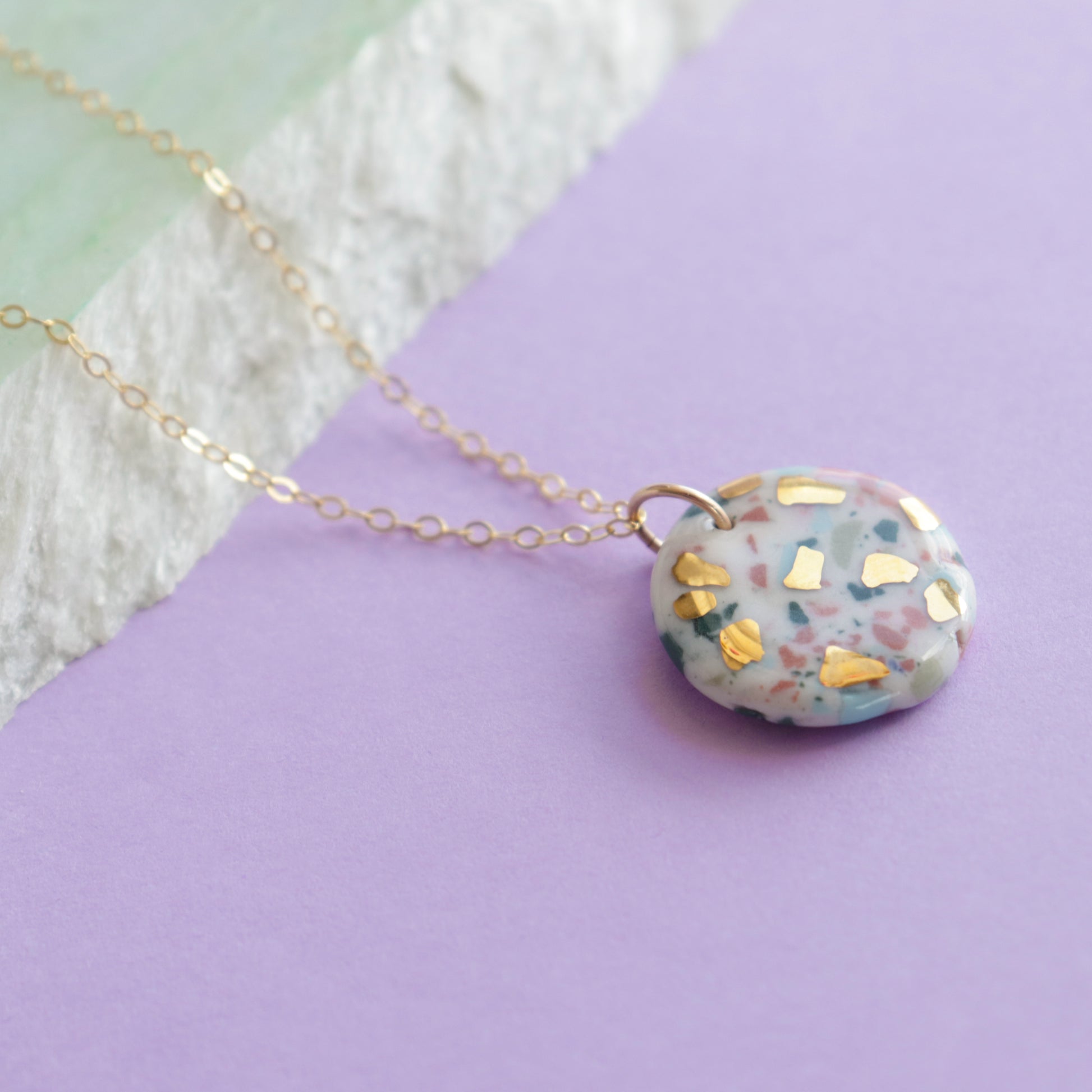 The Astilla Recycled Necklace by Rozenthal Jewelry features a round, pastel porcelain pendant with gold accents on a delicate gold chain, displayed against a lavender backdrop with textured white and green surfaces.
