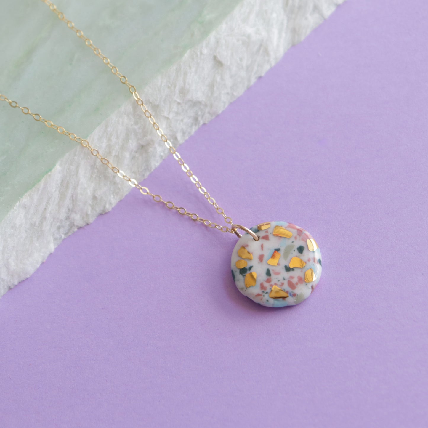 The Astilla Recycled Necklace by Rozenthal Jewelry features a round terrazzo-style pendant with colorful flecks on a delicate gold chain, displayed on a pastel green and purple background—an elegant, eco-friendly piece for daily wear.