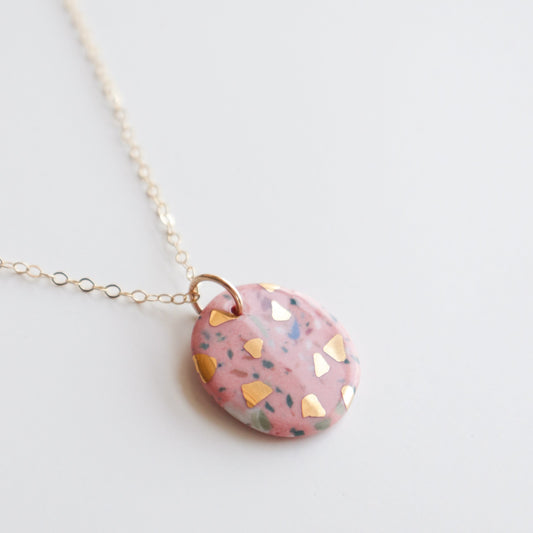The Astilla Recycled Necklace by Rozenthal Jewelry features a gold chain and a round pink porcelain pendant with gold and dark speckled accents—a stunning piece of handmade jewelry displayed on a white background.