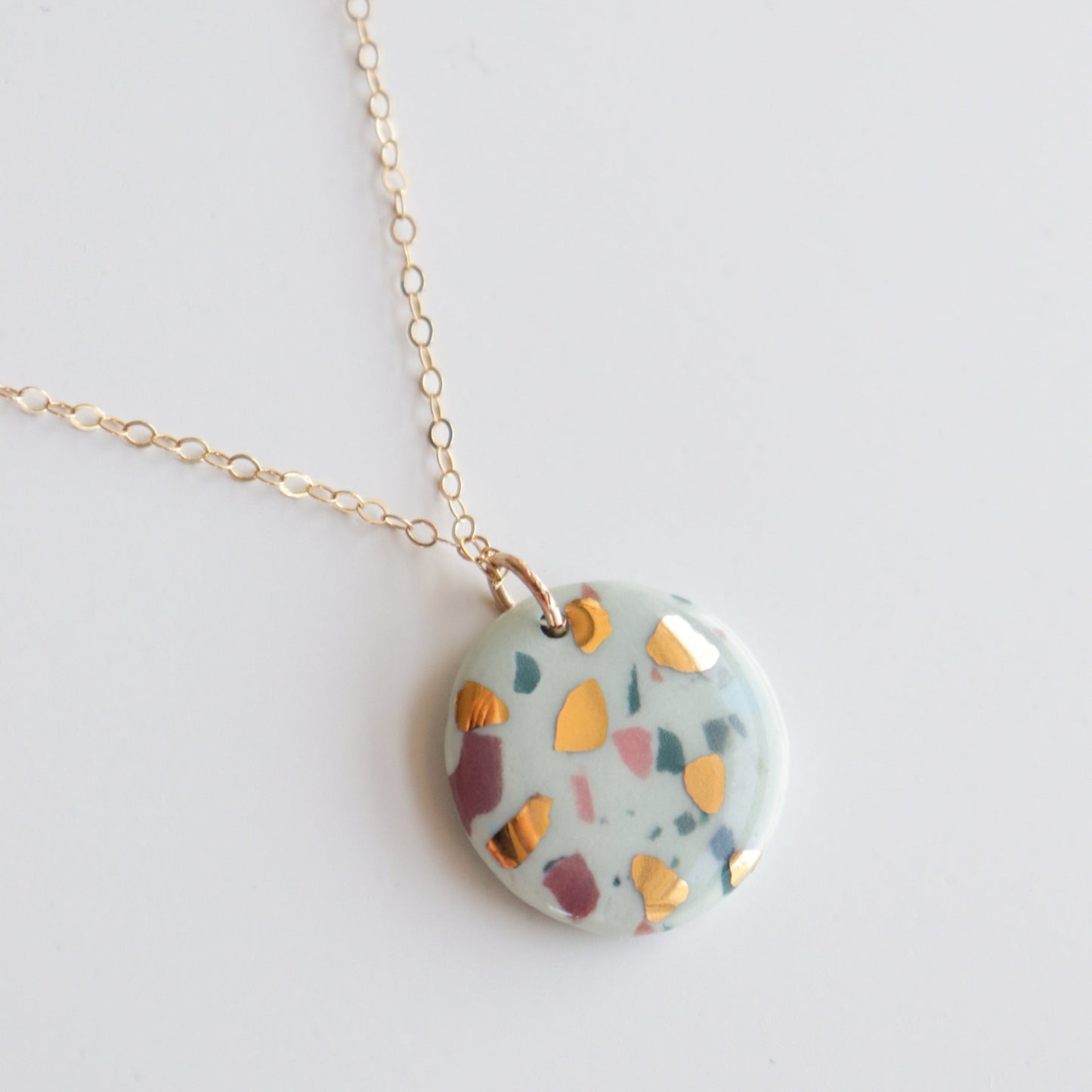 The Astilla Recycled Necklace by Rozenthal Jewelry features a gold chain with a round, glossy pendant showcasing a terrazzo-inspired pattern of colorful abstract shapes—yellow, pink, and green—on a white background.