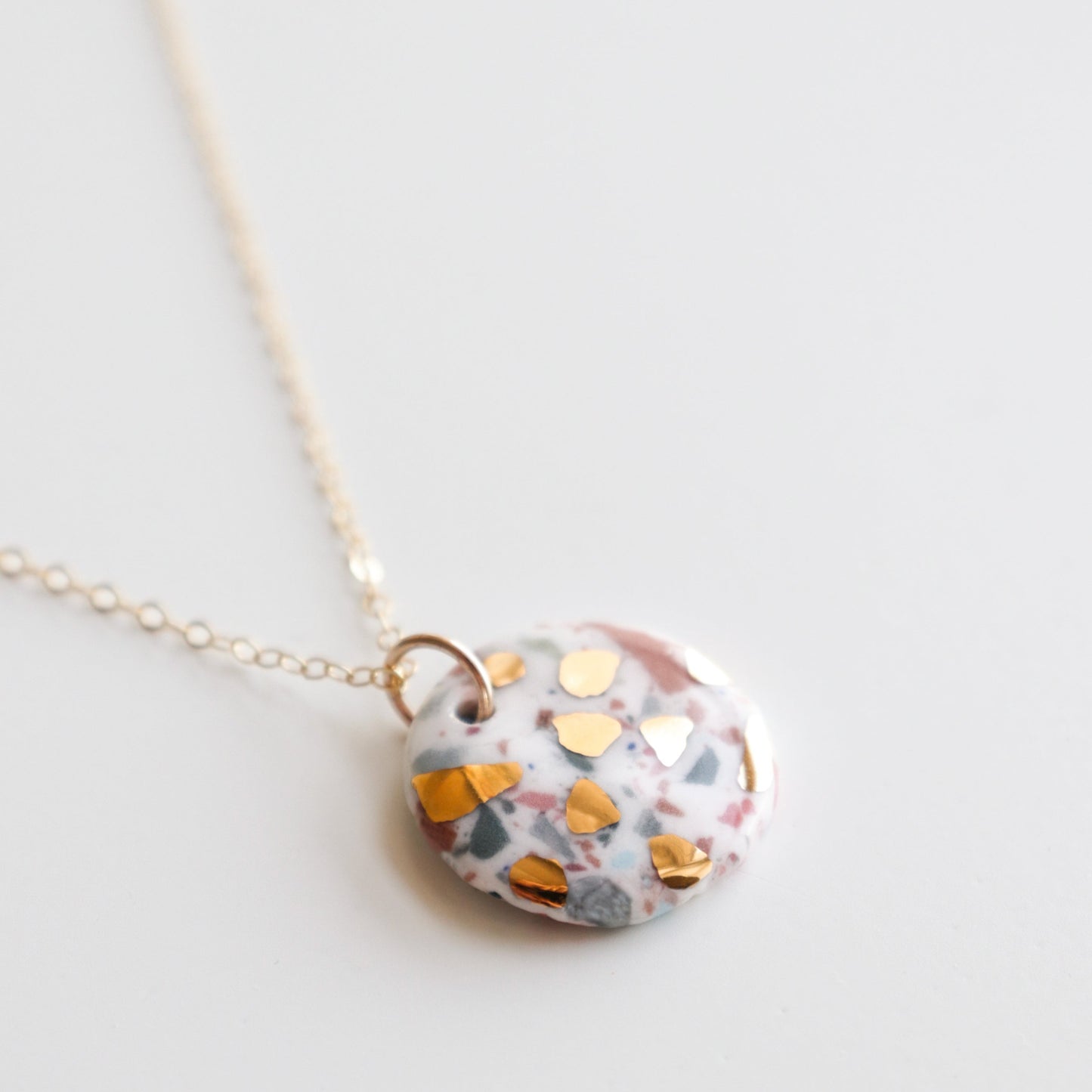 The Astilla Recycled Necklace by Rozenthal Jewelry features a delicate gold chain and a round pendant with a white base, colorful speckles, and gold foil accents, shown on a plain white background.