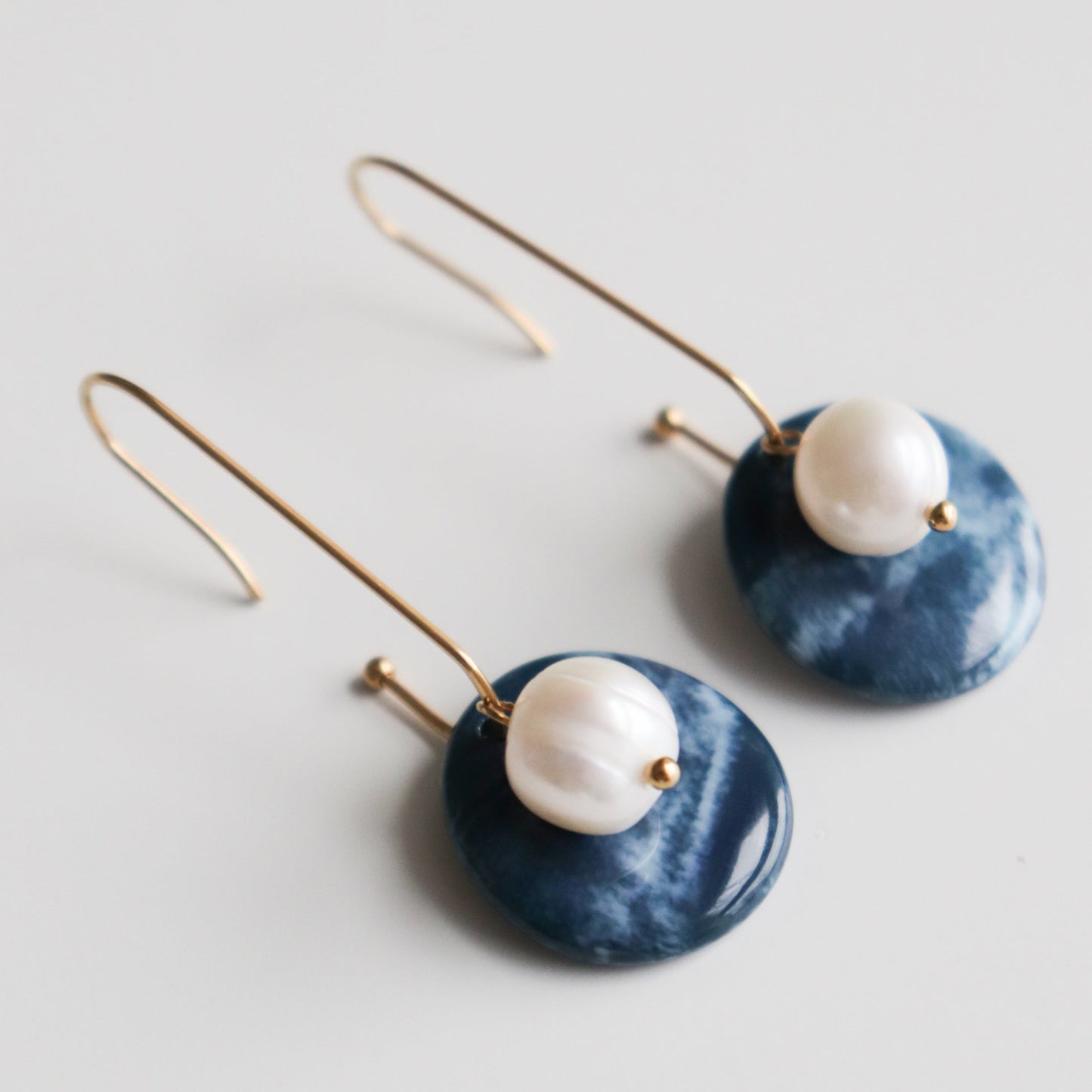 The Marée Earrings by Rozenthal Jewelry are elegant, hypoallergenic designer pieces with long gold hooks, round white pearls, and flat blue marble-like discs beneath the pearls, shown on a white background.
