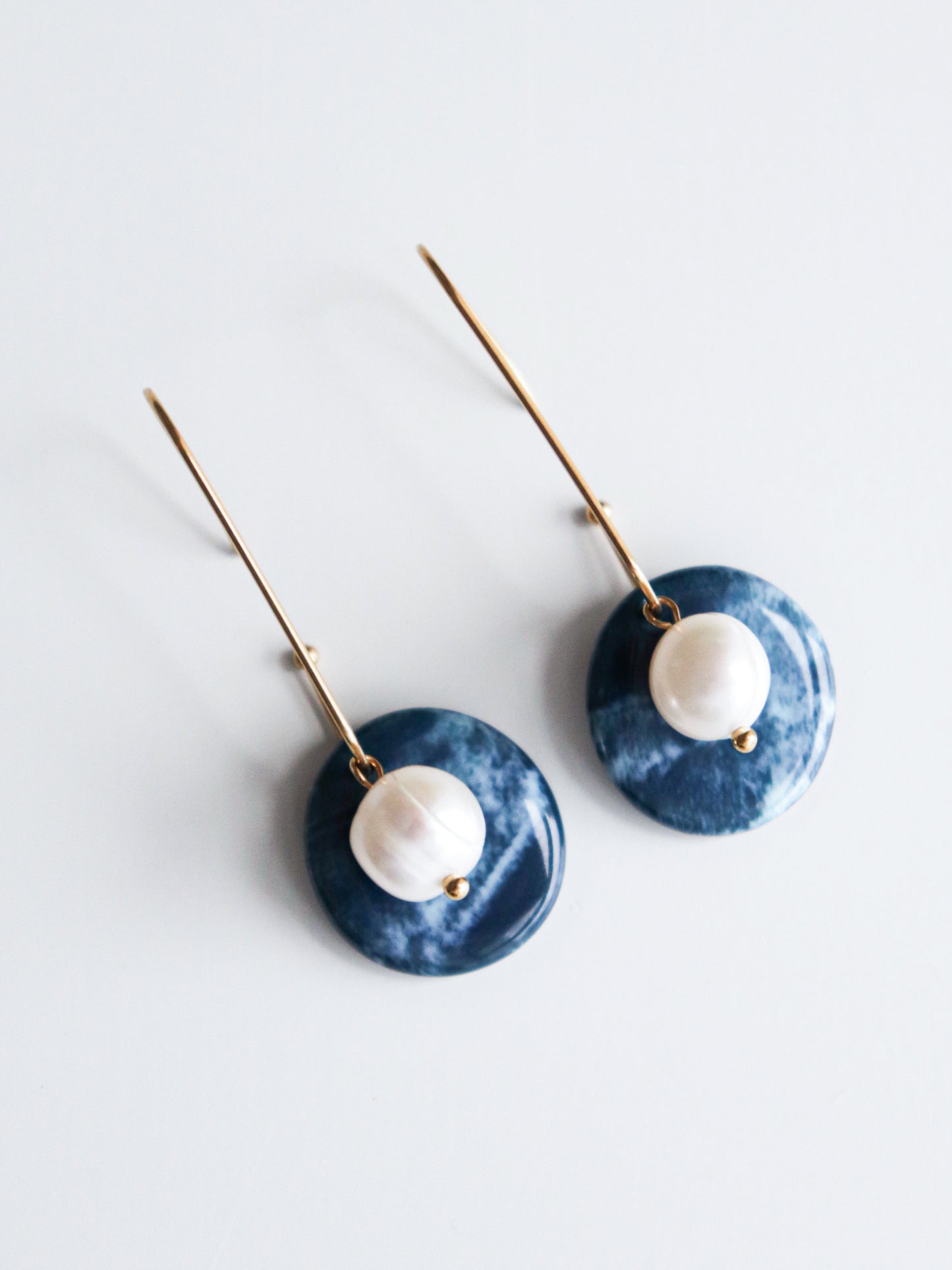 The Marée Earrings by Rozenthal Jewelry feature handmade gold hooks with round dark blue discs and a central white pearl bead. Lightweight and waterproof, these earrings are shown against a white background.