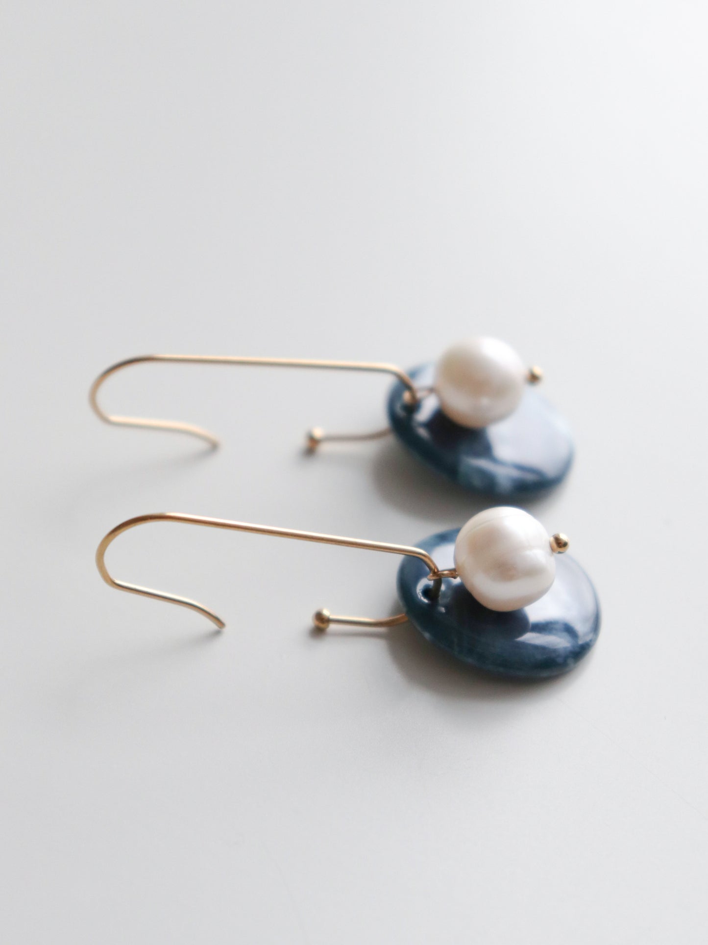 The Marée Earrings by Rozenthal Jewelry feature dangling gold hooks with irregular white pearls on round, dark blue porcelain discs—a handmade, refined piece for any porcelain jewelry collection. Shown on a light background.