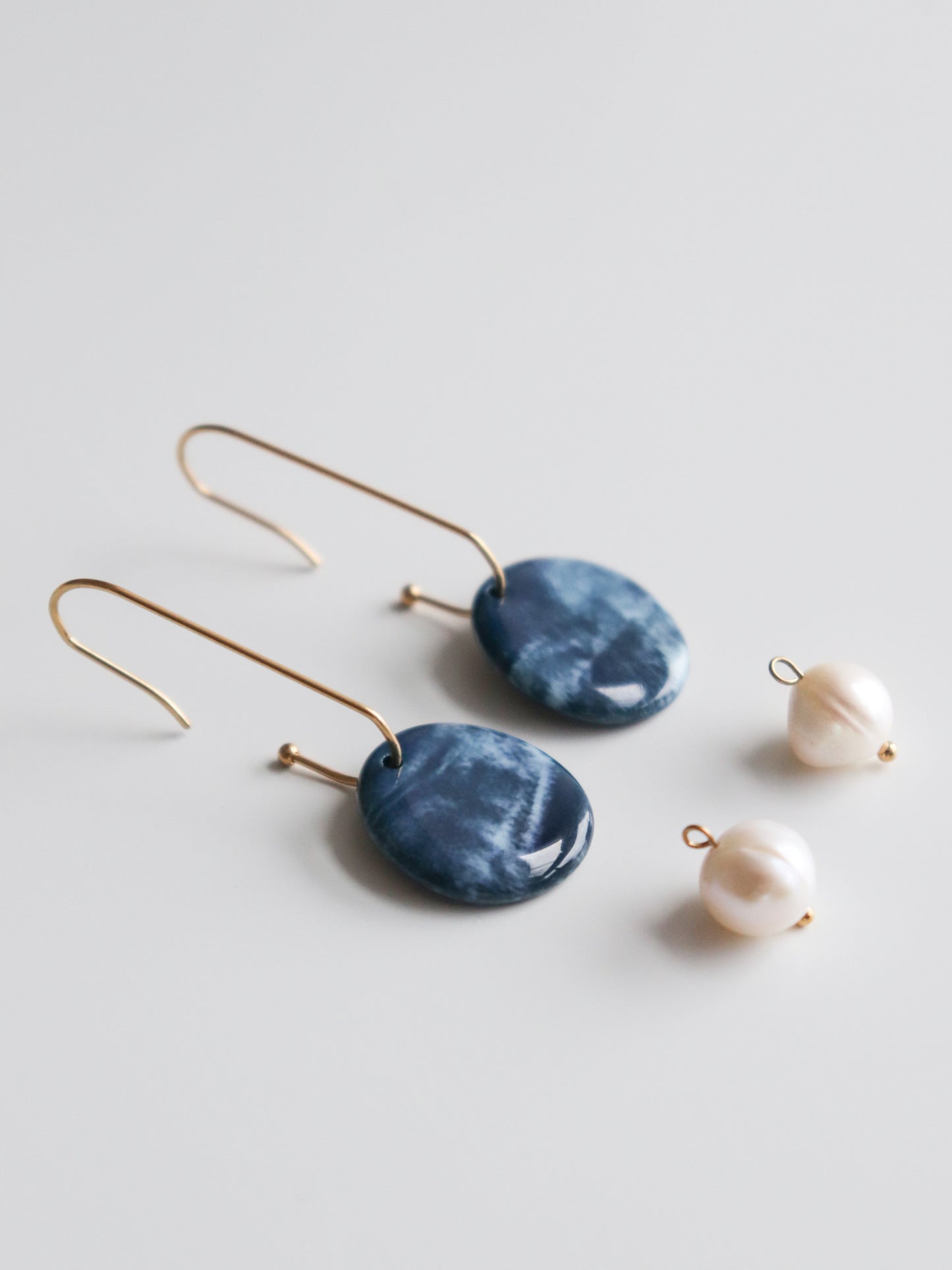 The Marée Earrings by Rozenthal Jewelry feature gold hooks and oval blue ceramic pendants, displayed on a white surface beside two loose round white pearls.