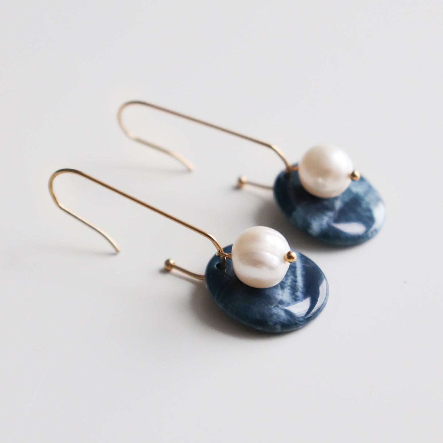 The Marée Earrings by Rozenthal Jewelry are gold hook earrings with white pearls and round dark blue marbled pendants. Hypoallergenic, lightweight, and waterproof, they are displayed on a white background.