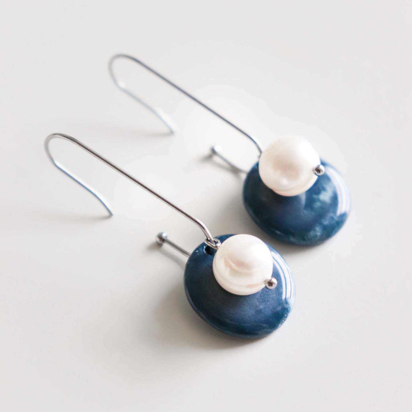 The Marée Earrings by Rozenthal Jewelry are lightweight, waterproof earrings with curved silver hooks, round dark blue discs, and white pearl-like beads on each earring, shown against a white background.