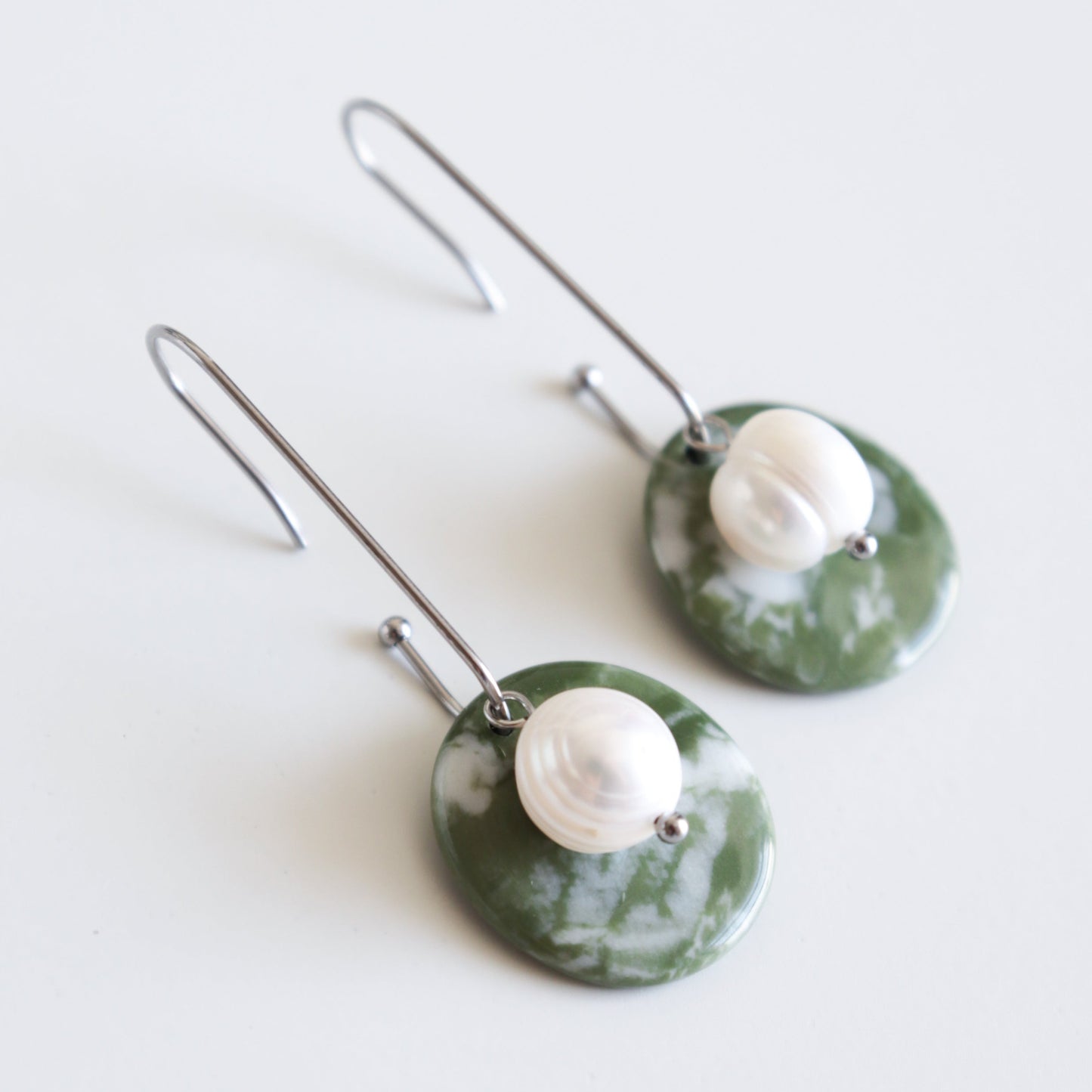 Marée Earrings by Rozenthal Jewelry: Hypoallergenic designer earrings with long silver hooks, each featuring a round green stone pendant topped with a white pearl bead. Handmade in Latvia and shown on a white background.