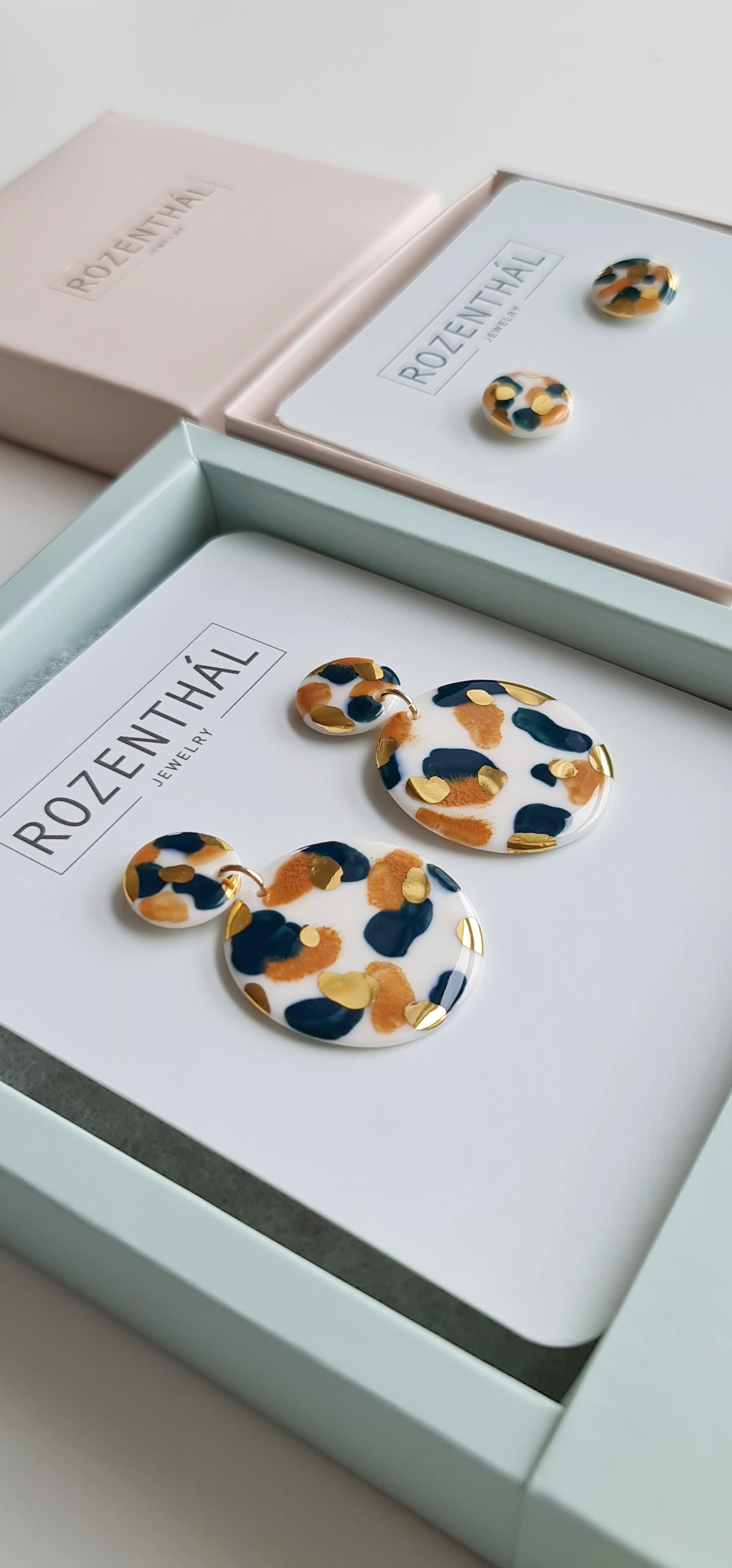 A close-up of two pairs of Tache Earrings by Rozenthal Jewelry—hand-painted porcelain with 24K gold, navy, and black leopard-print patterns—hypoallergenic, displayed in pastel boxes labeled ROZENTHAL JEWELRY.