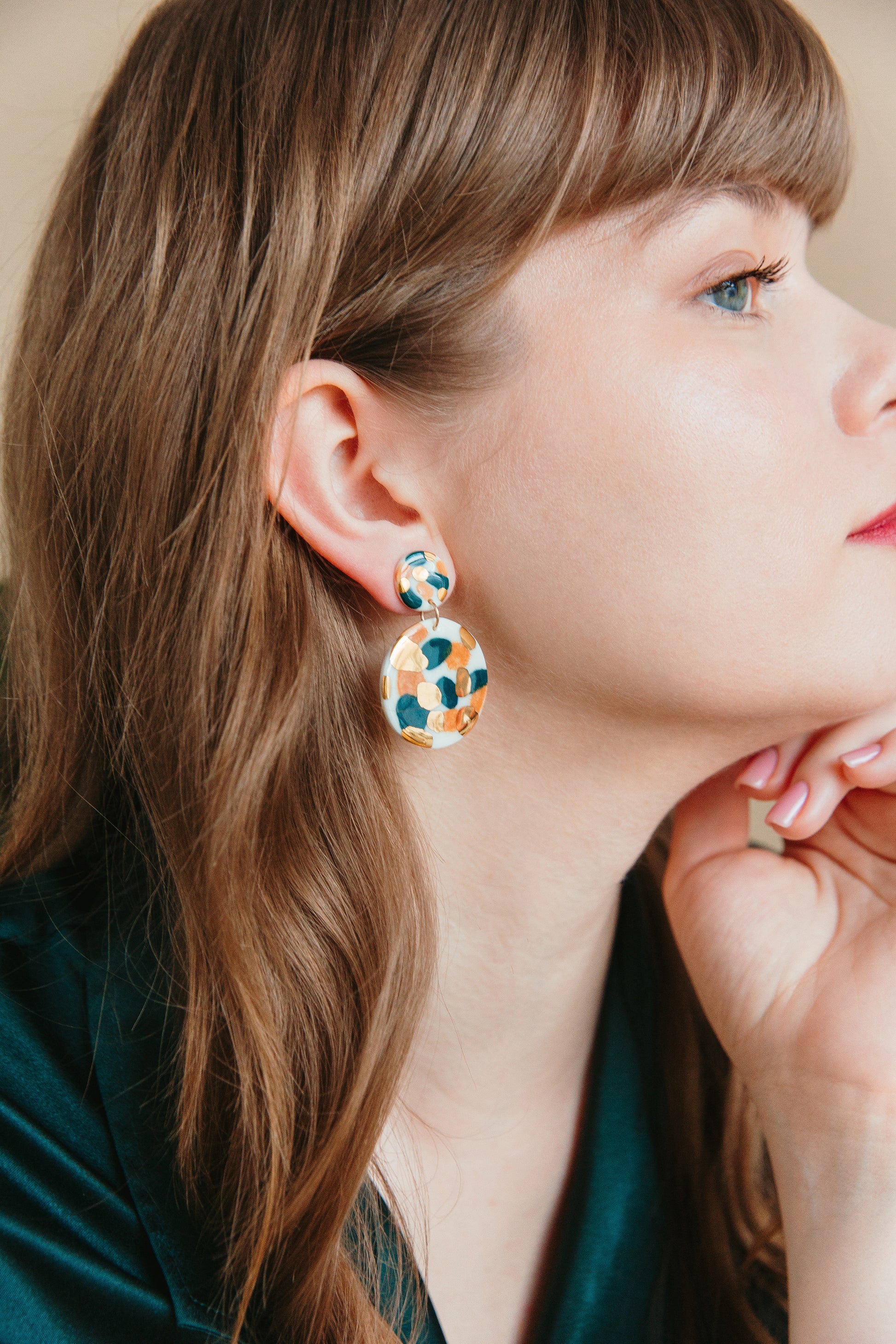 A woman with light brown hair and fair skin, shown in profile wearing a dark green blouse, rests her chin on her hand as she models Rozenthal Jewelrys Tache Earrings—large, round porcelain pieces hand-painted with 24K gold.