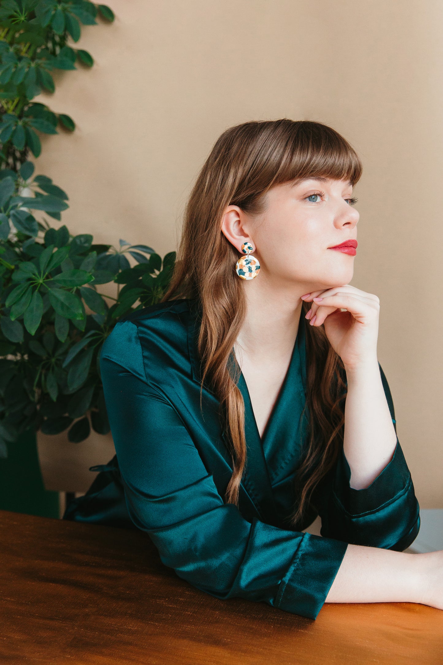A woman with long brown hair and red lipstick sits at a wooden table, wearing a dark green satin blouse and Rozenthal Jewelry’s Tache Earrings. A leafy plant and beige wall provide the background.