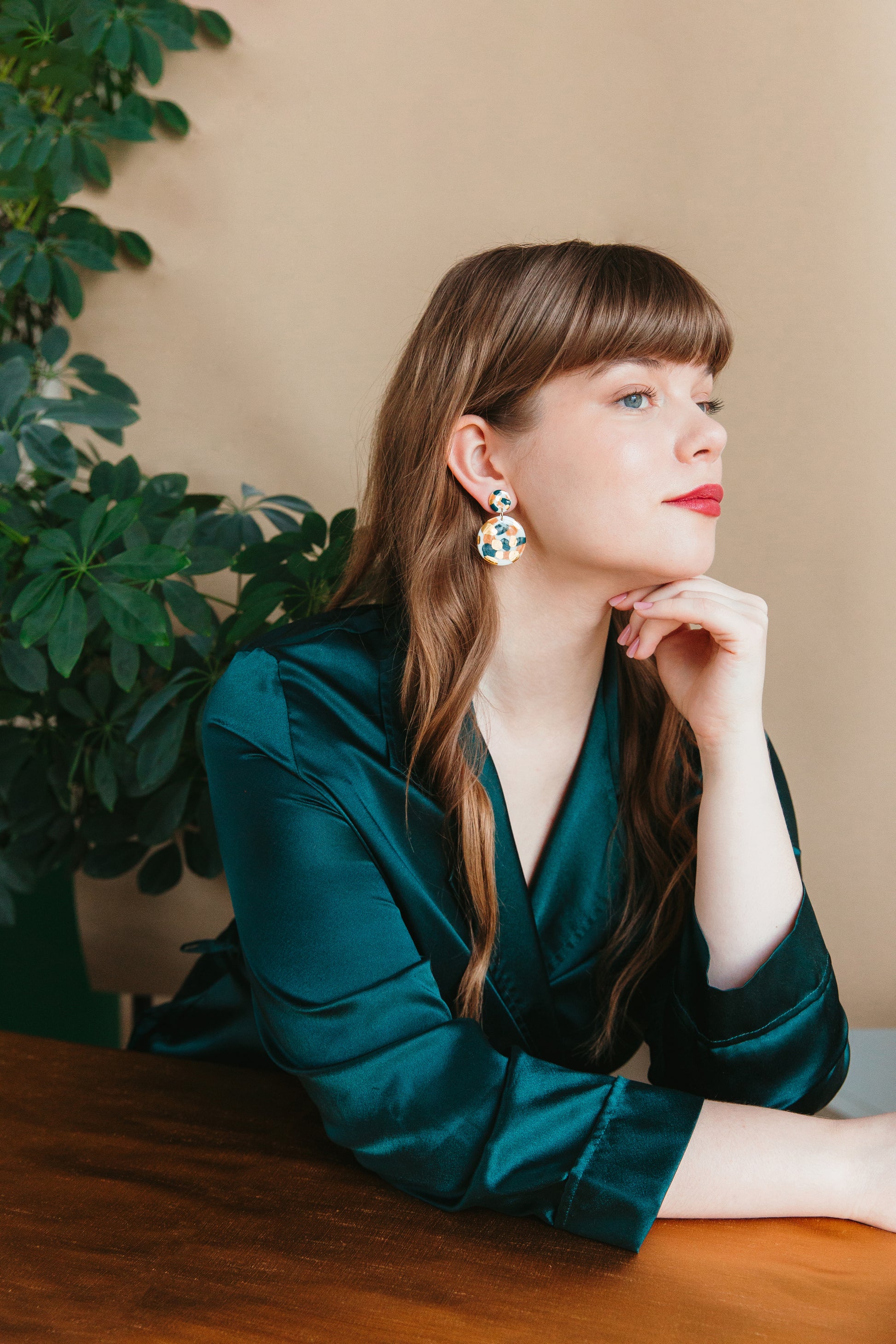 A woman with long brown hair and red lipstick sits at a wooden table, wearing a dark green satin blouse and Rozenthal Jewelry’s Tache Earrings. A leafy plant and beige wall provide the background.