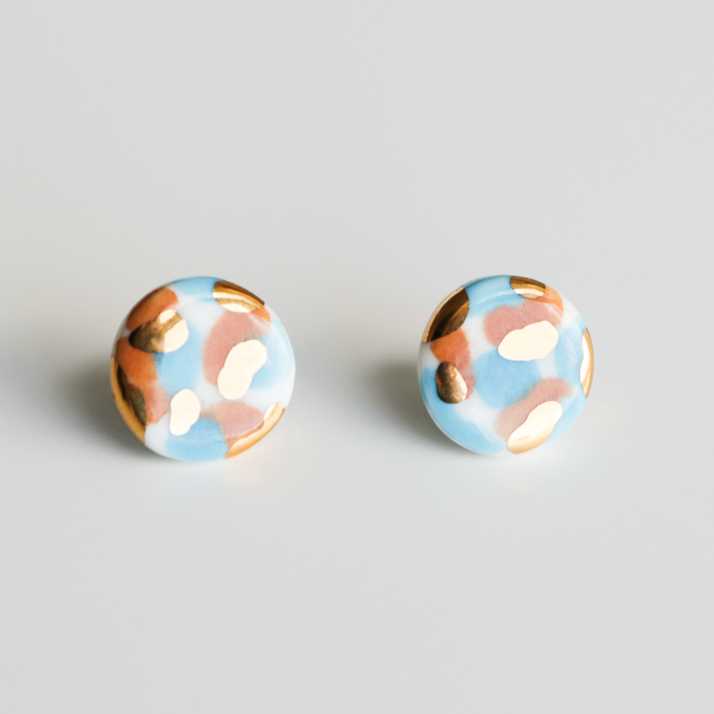 The Tache Studs / S by Rozenthal Jewelry are round porcelain earrings with a blue base, featuring hand-painted gold, white, and brown spots. Photographed on a plain white background.