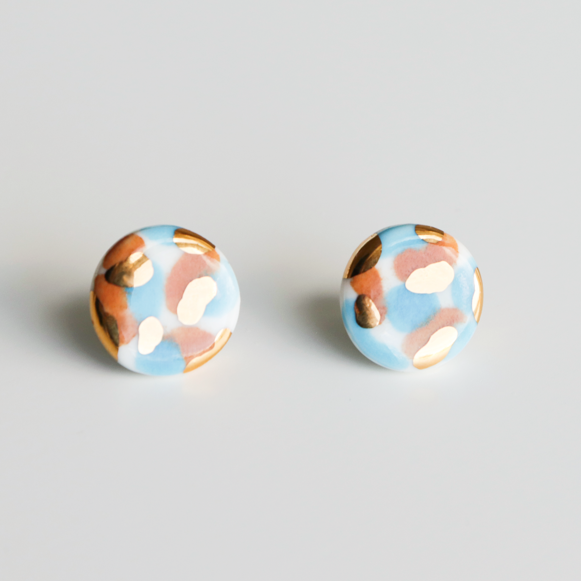 The Tache Studs / S by Rozenthal Jewelry are round porcelain earrings with a blue base, featuring hand-painted gold, white, and brown spots. Photographed on a plain white background.