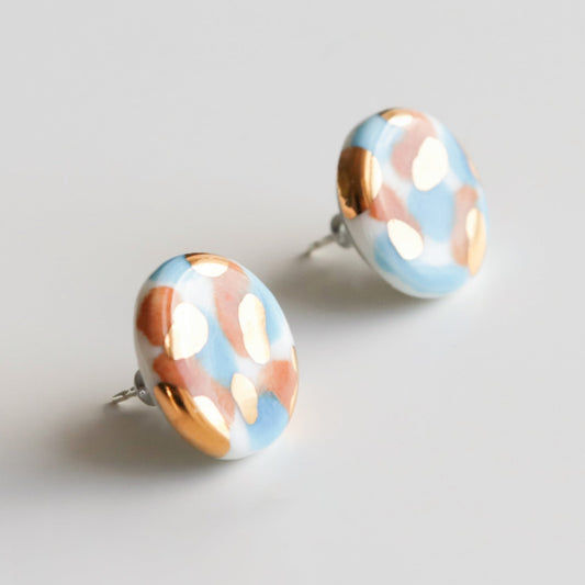 The Tache Studs / S by Rozenthal Jewelry are lightweight, waterproof porcelain statement earrings with a glossy finish and an abstract pattern in light blue, white, and gold. Displayed on a plain light background.