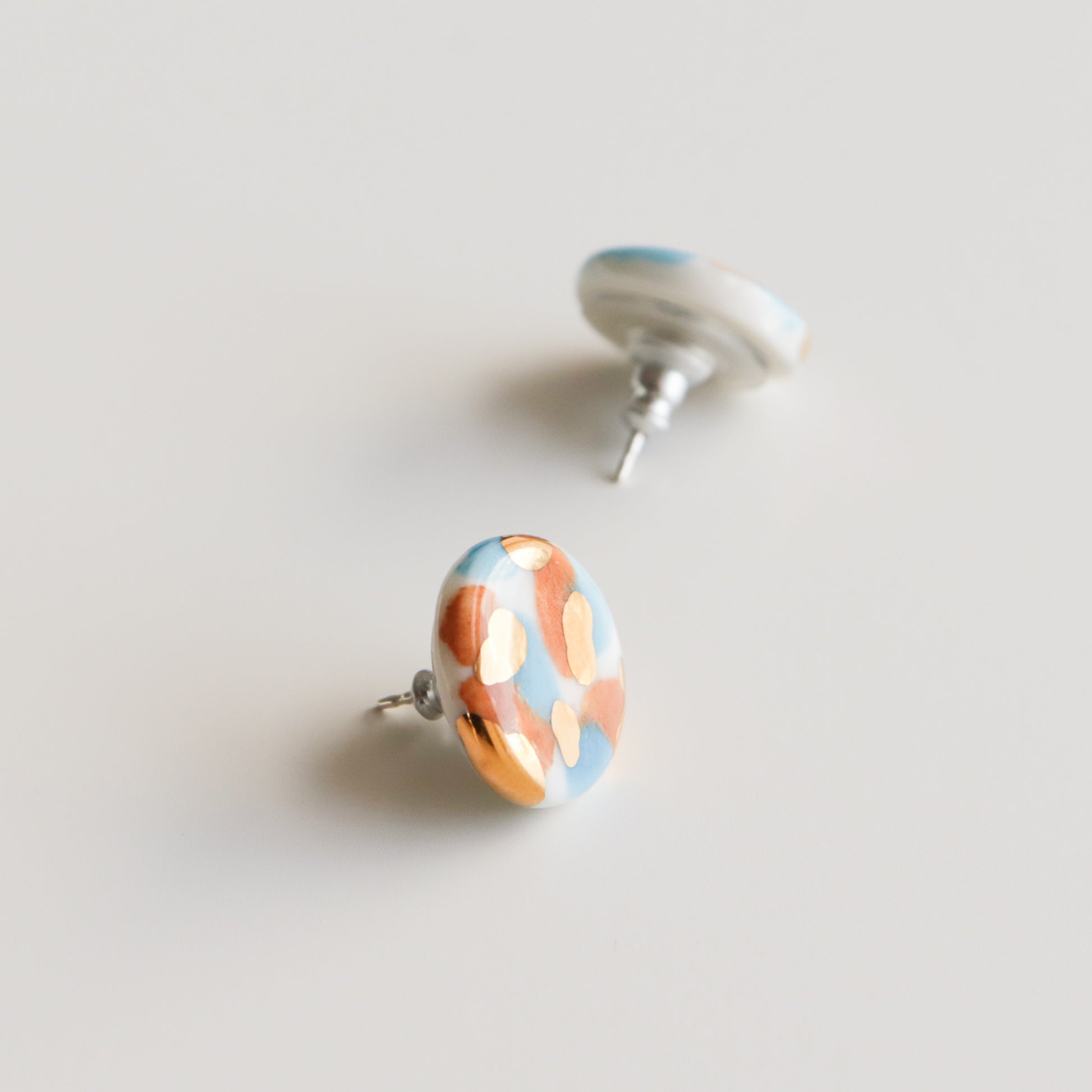 The Rozenthal Jewelry Tache Studs / S are round porcelain earrings with a glossy finish and an abstract blue, white, and copper pattern. These hypoallergenic sterling silver studs are shown on a plain white background.