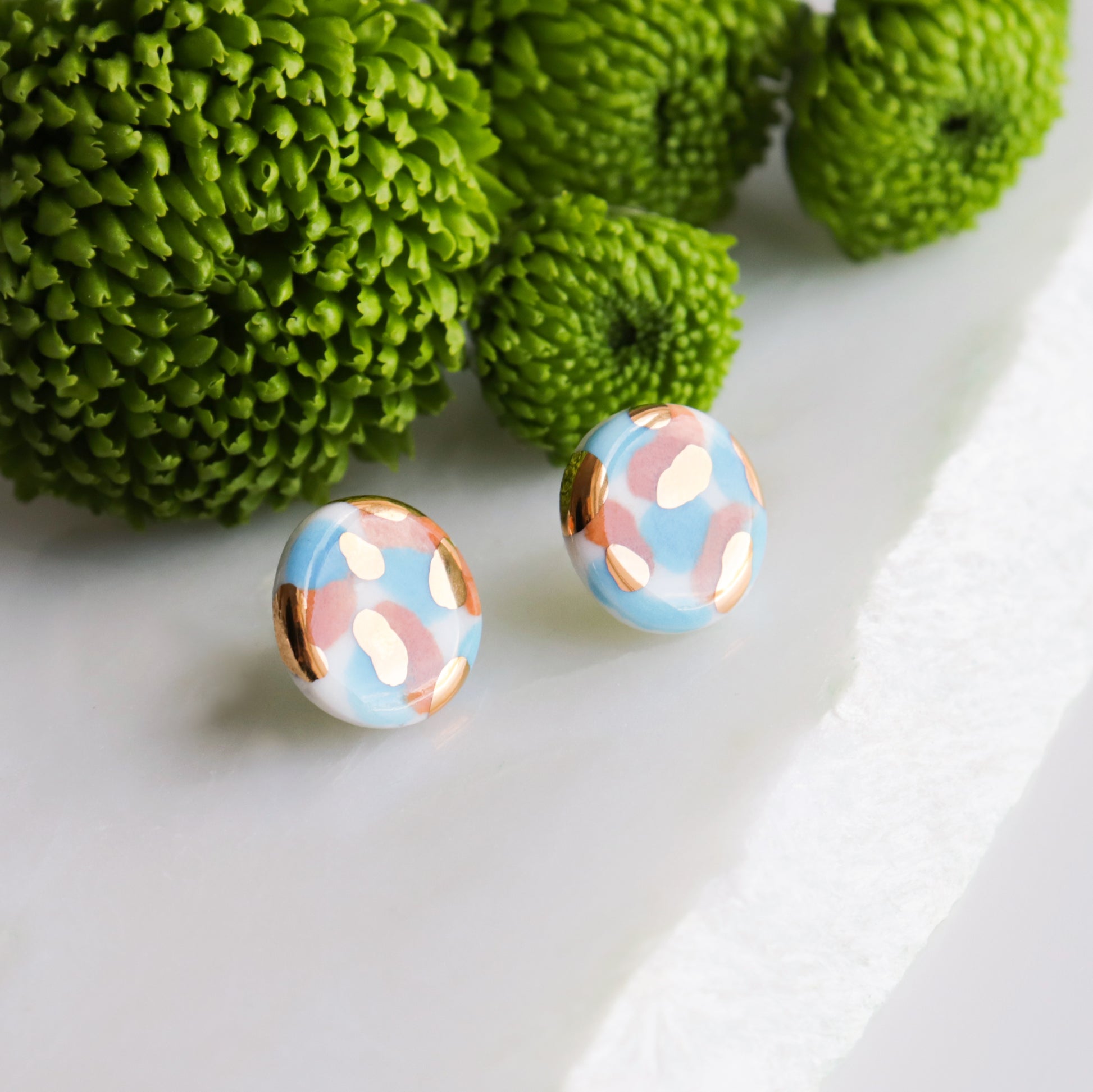 The Tache Studs / S by Rozenthal Jewelry are round porcelain earrings with a blue, white, and copper abstract pattern, displayed on a white surface beside green textured flowers—a stunning hand-painted jewelry piece.