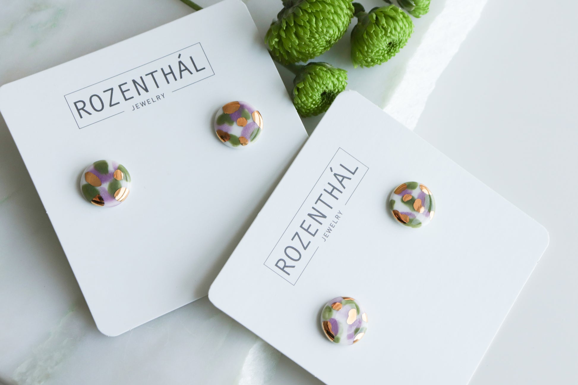 Two pairs of Tache Studs / S by Rozenthal Jewelry—colorful, patterned, and handmade in Latvia—are displayed on branded cards atop a white surface next to green decorative flowers.