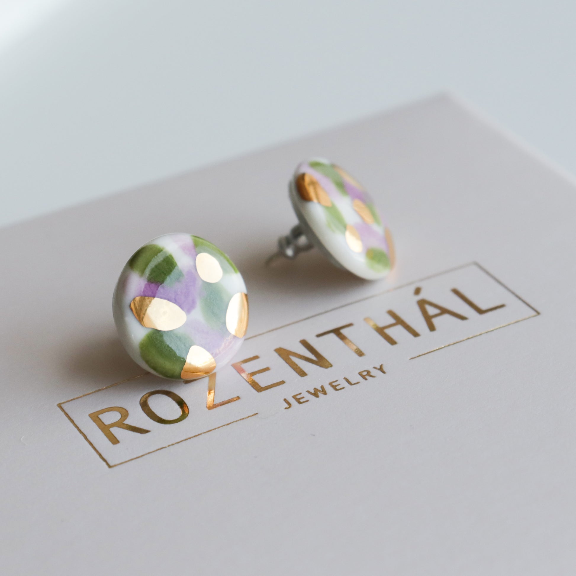 The Tache Studs / S by Rozenthal Jewelry are round ceramic earrings featuring green, lavender, and gold abstract patterns. Lightweight and waterproof, they come displayed on a white card with gold-embossed branding.