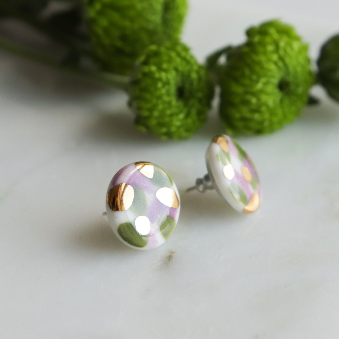 The Tache Studs / S by Rozenthal Jewelry are round, lightweight, waterproof earrings featuring a white base with green and light purple patterns and gold accents. Shown on marble with green flowers in the background.