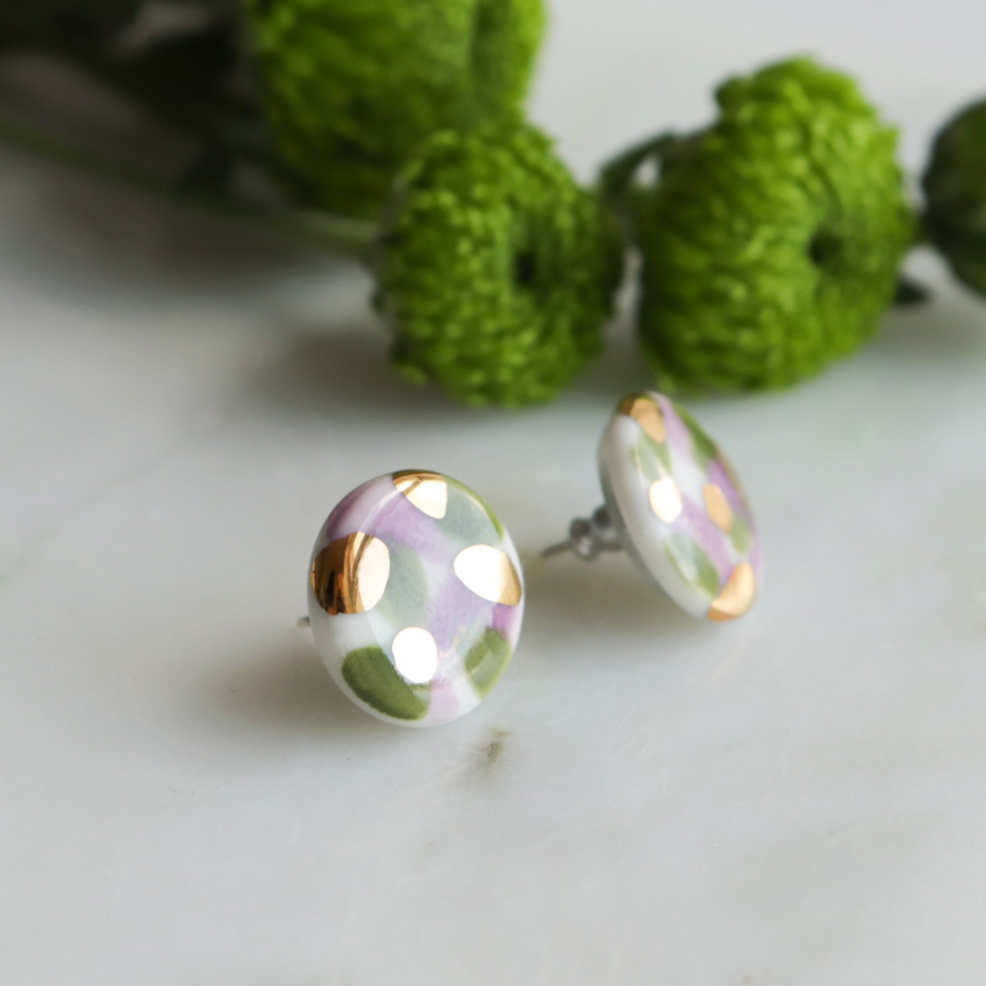 The Tache Studs / S by Rozenthal Jewelry are round, lightweight, waterproof earrings featuring a white base with green and light purple patterns and gold accents. Shown on marble with green flowers in the background.