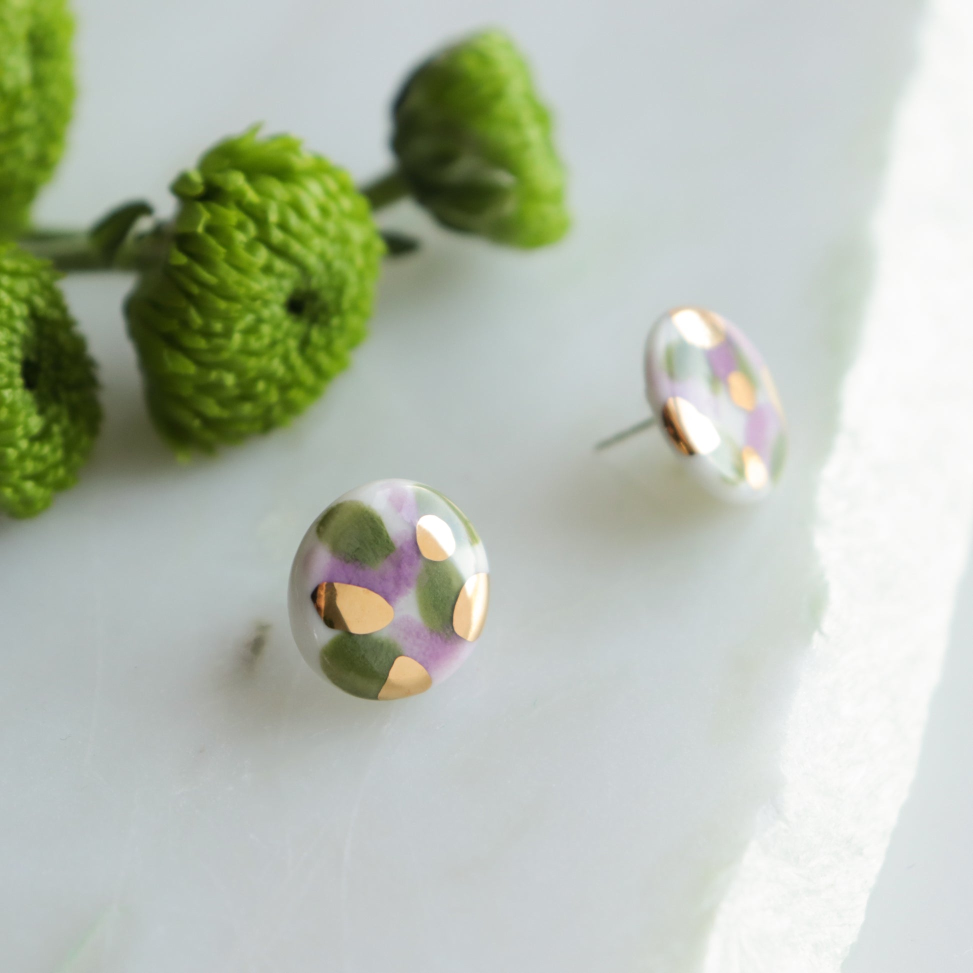 The Tache Studs / S by Rozenthal Jewelry are round handmade earrings featuring gold, olive green, and lilac spots on white, shown on marble next to green flowers.