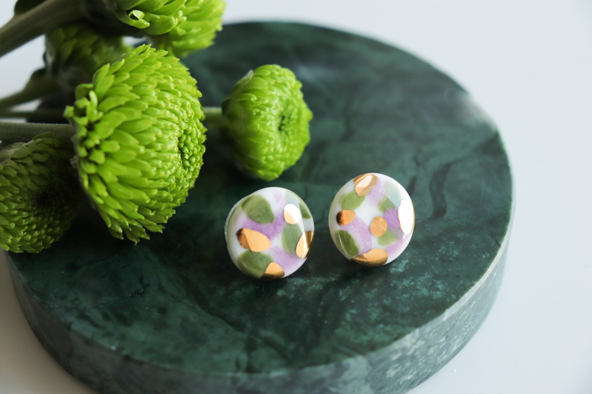 The Tache Studs / S by Rozenthal Jewelry—round, colorful handmade earrings with abstract patterns and gold accents—are displayed on a dark green marble surface beside bright green flowers.