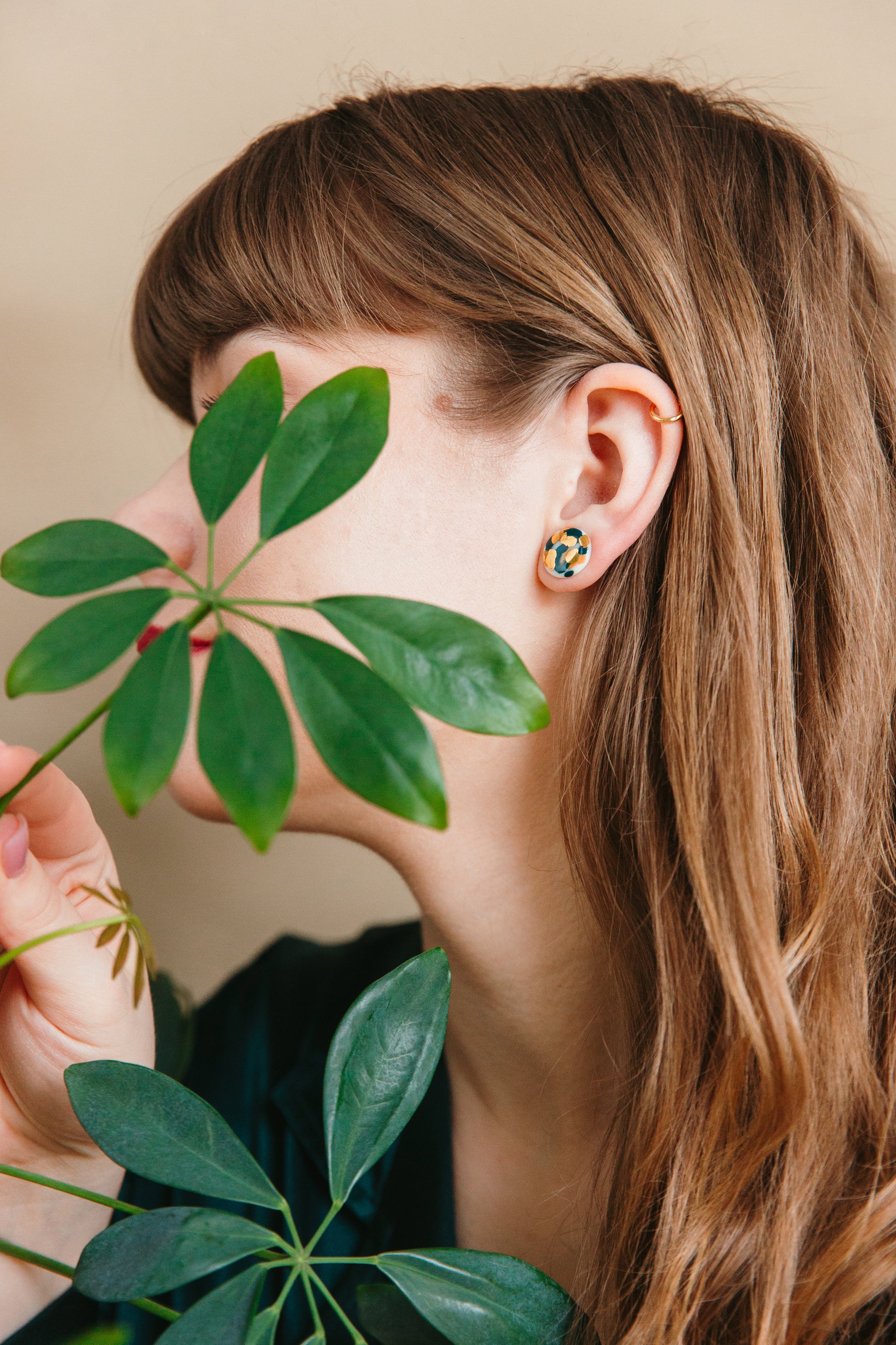 A woman with long brown hair faces sideways, partially hidden by green leaves. She wears Rozenthal Jewelrys Tache Studs / S and a dark top, with her ear and part of her face visible through the foliage.