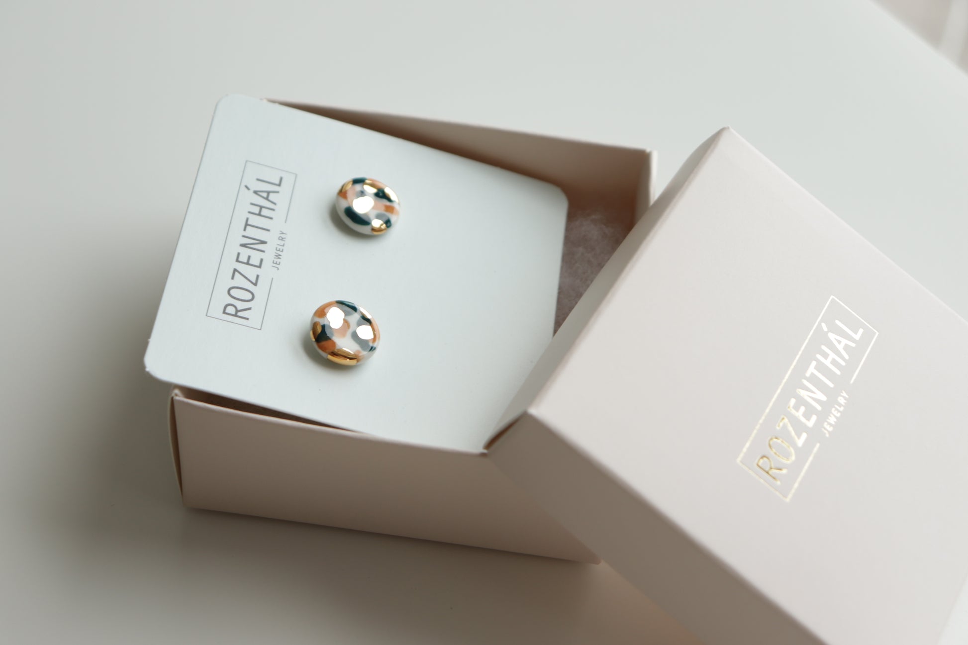 A pair of Tache Studs / S by Rozenthal Jewelry sits on a branded card inside an open pale pink Rozenthal Jewelry box, displayed on a white surface.