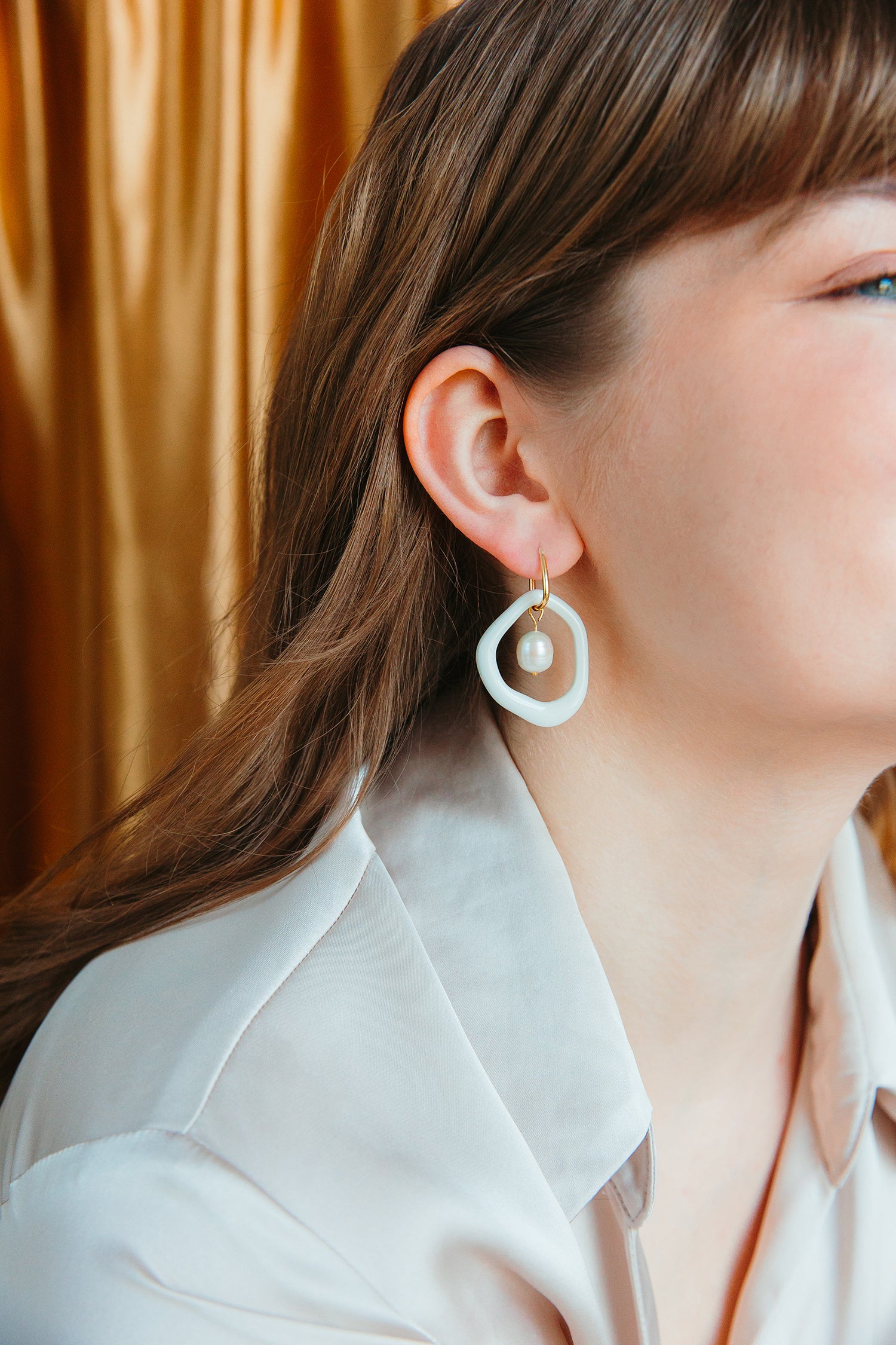 A woman with long brown hair wears a pale satin blouse and Rozenthal Jewelry’s Arp Pearl Hoops—white geometric porcelain earrings with dangling freshwater pearls—against a gold satin curtain, her face and shoulder in profile.