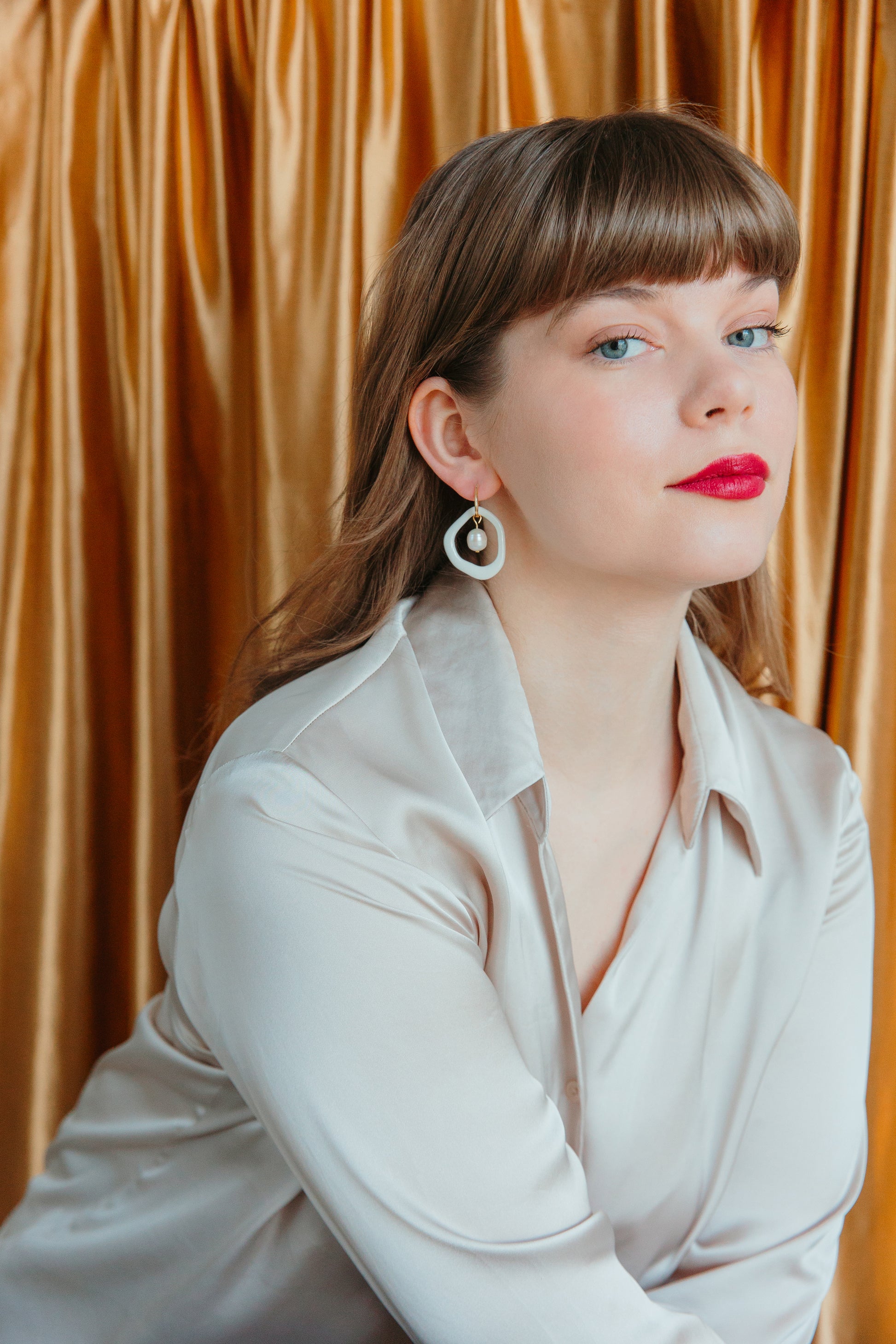 A woman with straight brown hair and red lipstick models Rozenthal Jewelrys Arp Pearl Hoops, handmade porcelain earrings, while wearing a light satin blouse in front of gold curtains.