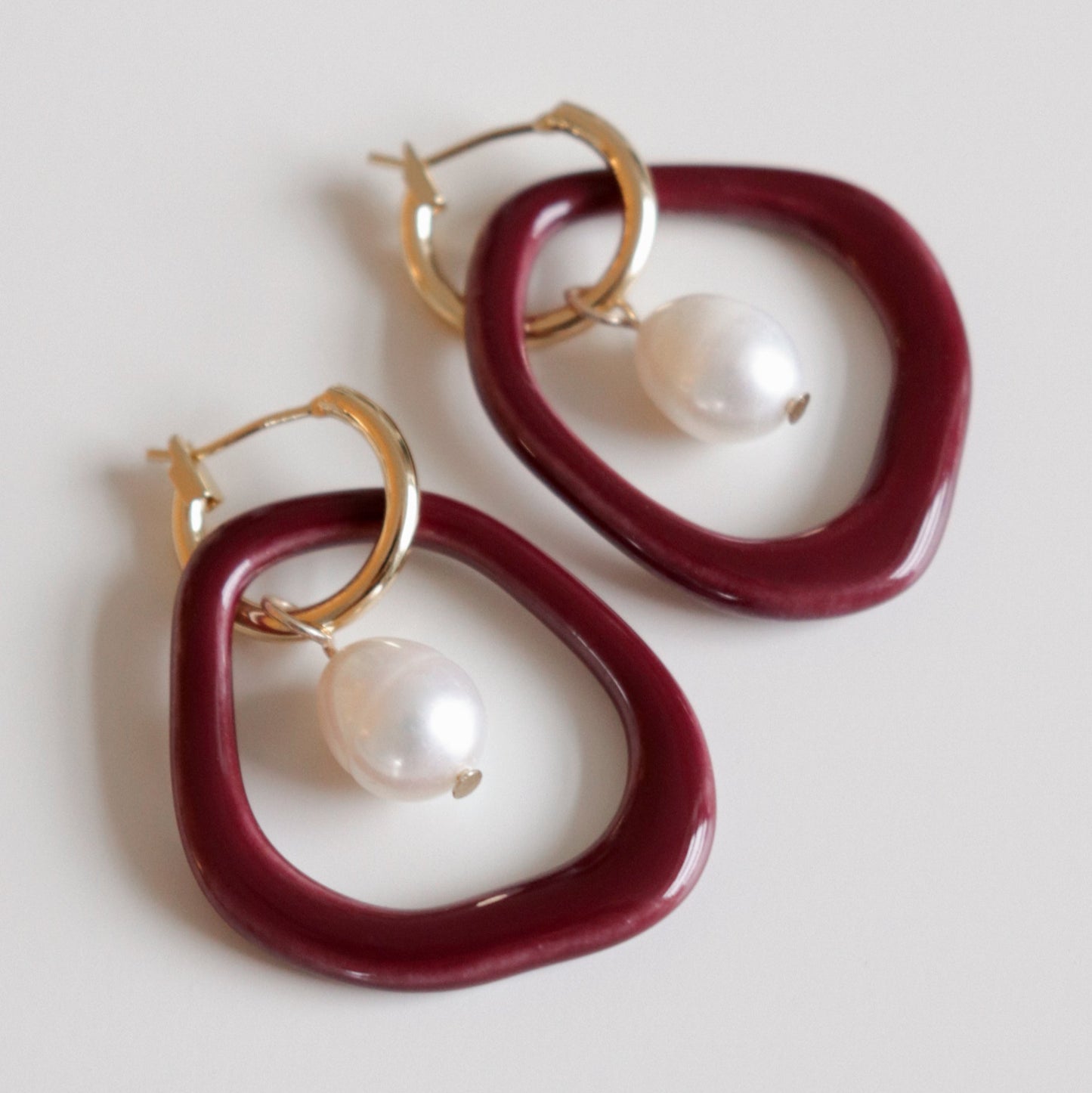 The Arp Pearl Hoops by Rozenthal Jewelry feature gold hoops with irregular maroon rings and dangling white freshwater pearls, ideal for fans of unique handmade jewelry, shown against a light background.