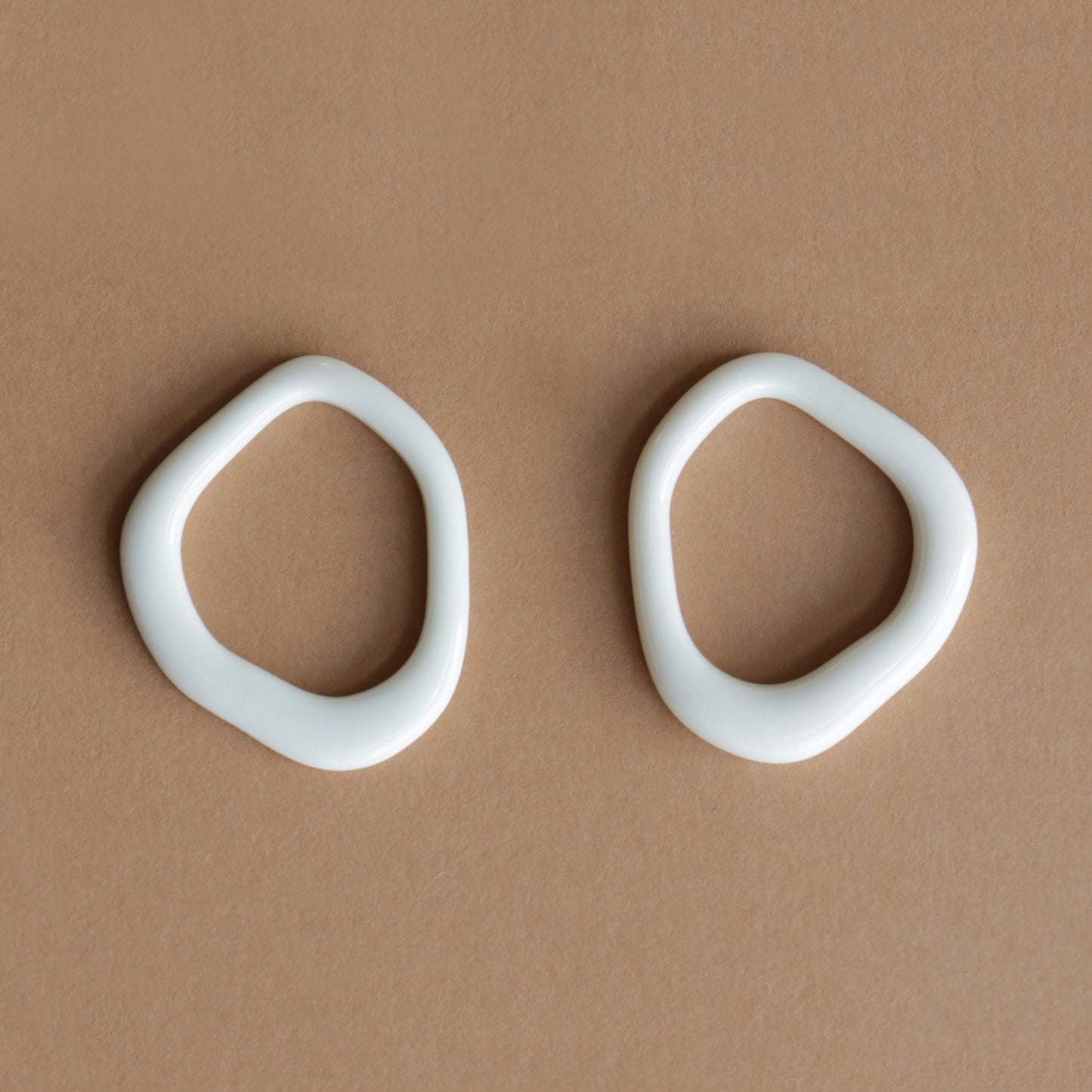 The Arp Pearl Hoops by Rozenthal Jewelry feature two irregular, white, oval ceramic rings with smooth edges and organic, asymmetrical forms, highlighting the unique charm of handmade jewelry on a light brown surface.