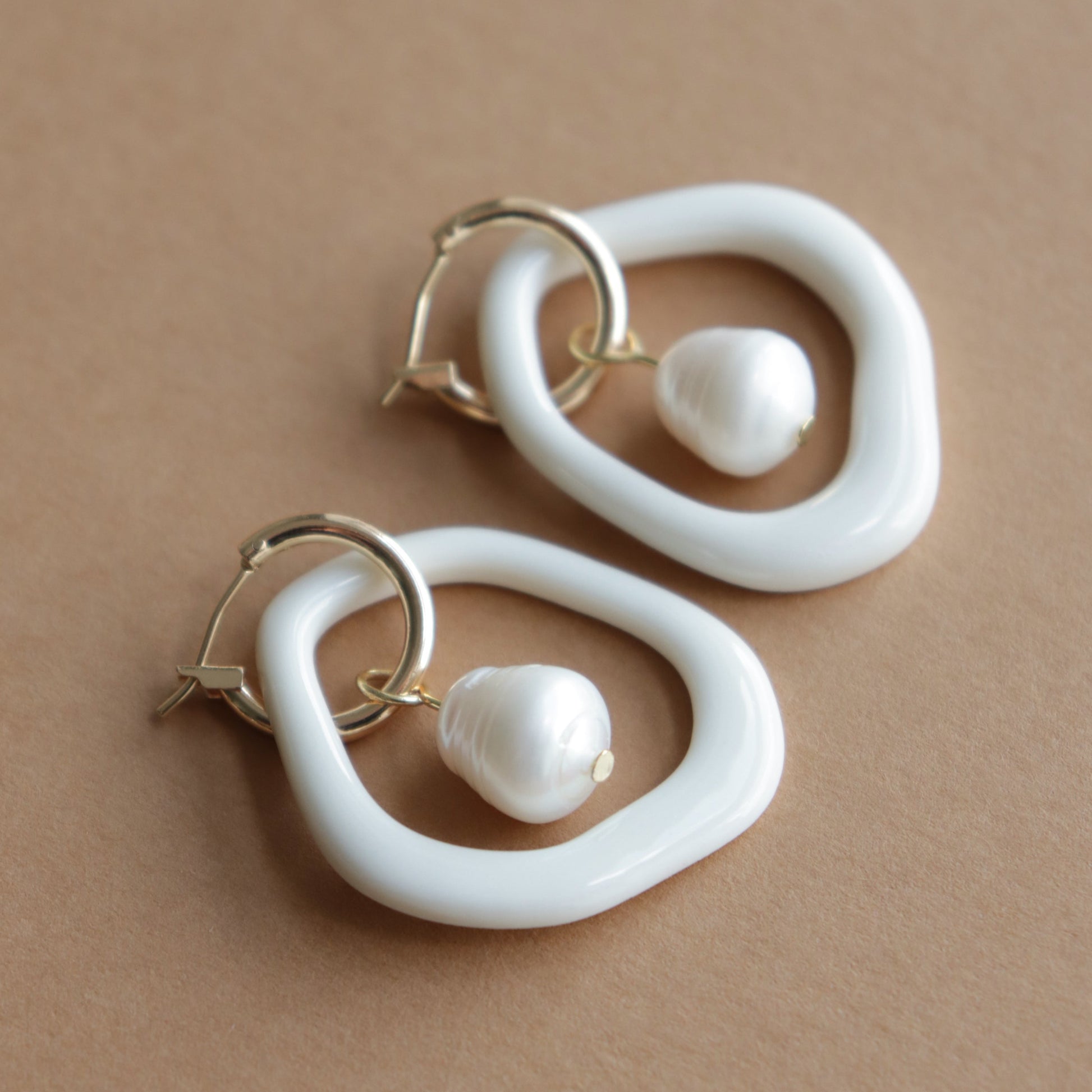 The Arp Pearl Hoops by Rozenthal Jewelry are gold hoop earrings with irregular white resin loops and dangling pearl-like beads, handmade to evoke the elegance of freshwater pearls. Shown on a beige background.