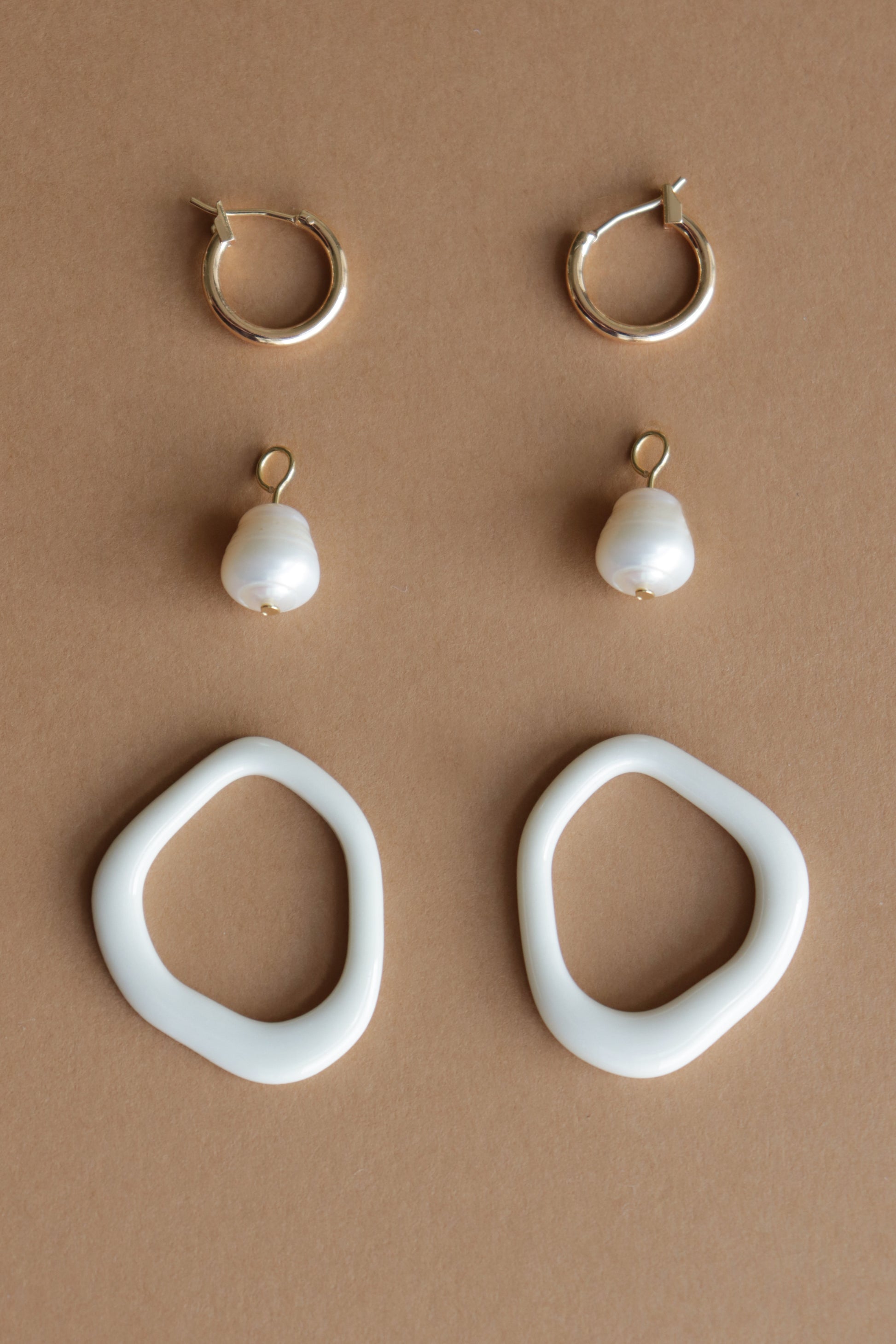 Three pairs of Rozenthal Jewelry earrings on a brown background: gold hoops on top, Arp Pearl Hoops with freshwater pearls in the middle, and large white porcelain hoops at the bottom.