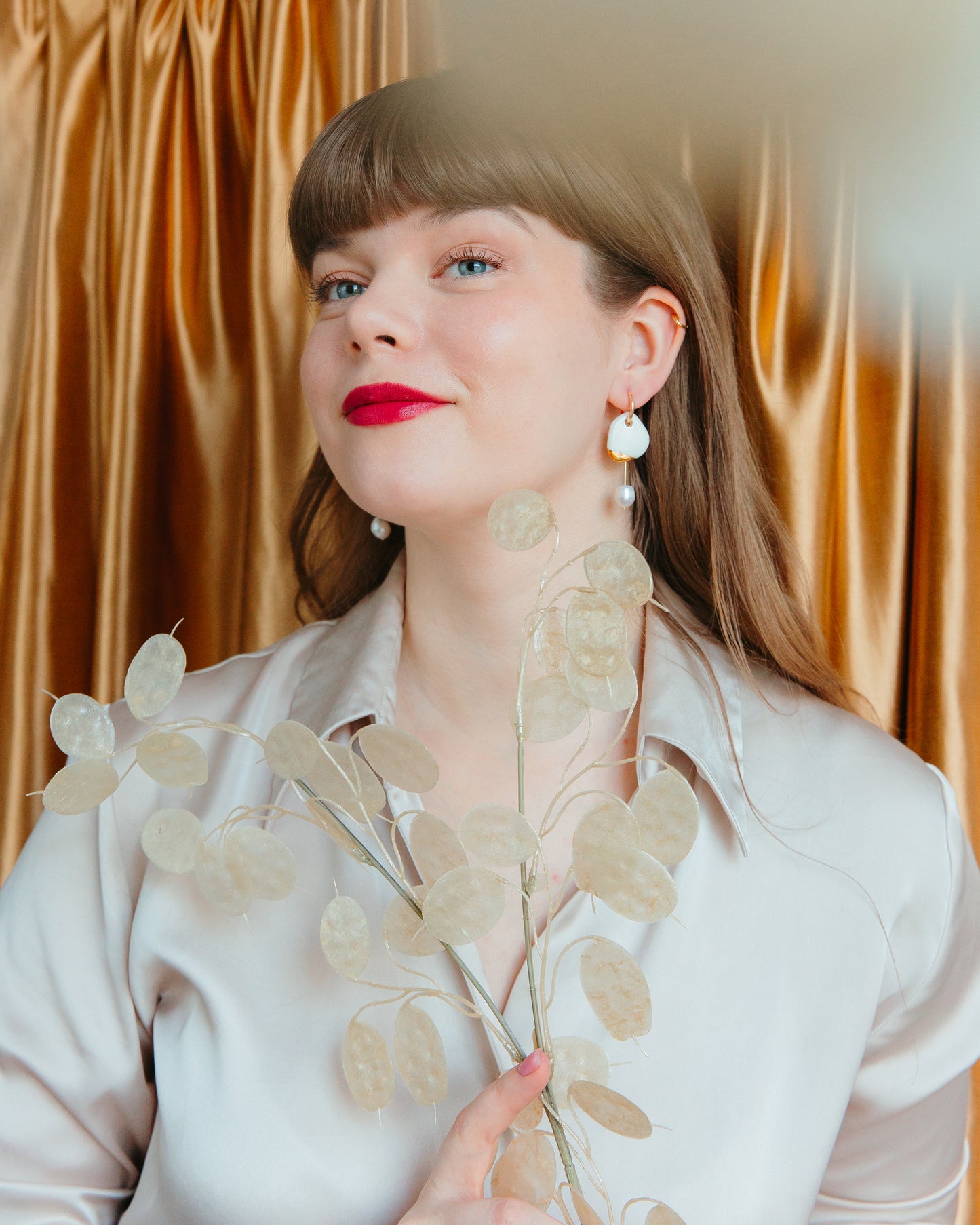 A woman with long brown hair and red lipstick stands before golden curtains, wearing Rozenthal Jewelrys Arp Pearl Hoops and a satiny beige blouse while holding delicate translucent seed pods.