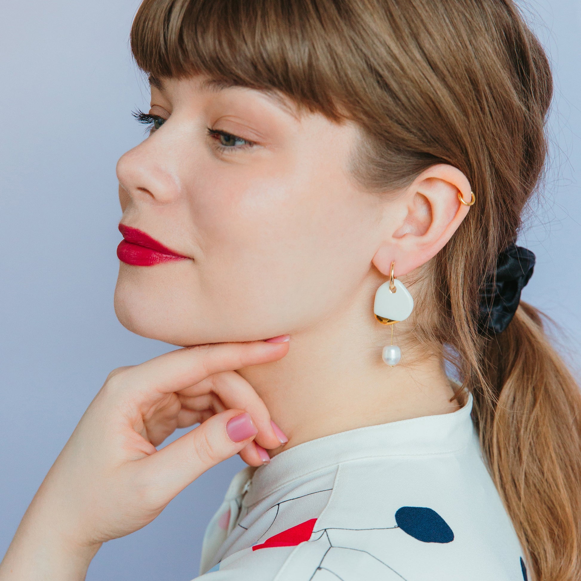 A woman with light brown hair in a ponytail wears red lipstick and Rozenthal Jewelrys Arp Pearl Hoops. She poses in a white top with red and navy patterns, hand under her chin, against a light purple background.