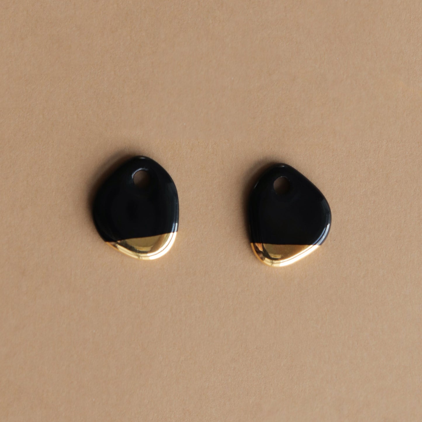 The Arp Pearl Hoops by Rozenthal Jewelry feature two irregularly shaped black and gold charms with a small top hole, ideal for handmade jewelry, shown on a light brown surface.