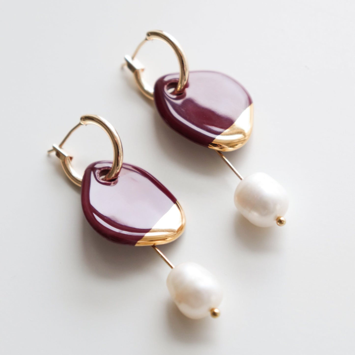 The Arp Pearl Hoops by Rozenthal Jewelry feature gold hooks, maroon teardrop accents with gold tips, and single freshwater pearls—an elegant pair of handmade earrings displayed on a white background.