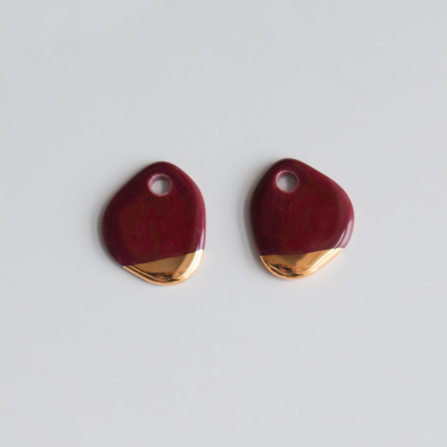 The Arp Pearl Hoops by Rozenthal Jewelry feature two maroon enamel charms with irregular shapes, small top holes, and gold accents at the bottom, capturing an artisanal feel against a plain white backdrop.