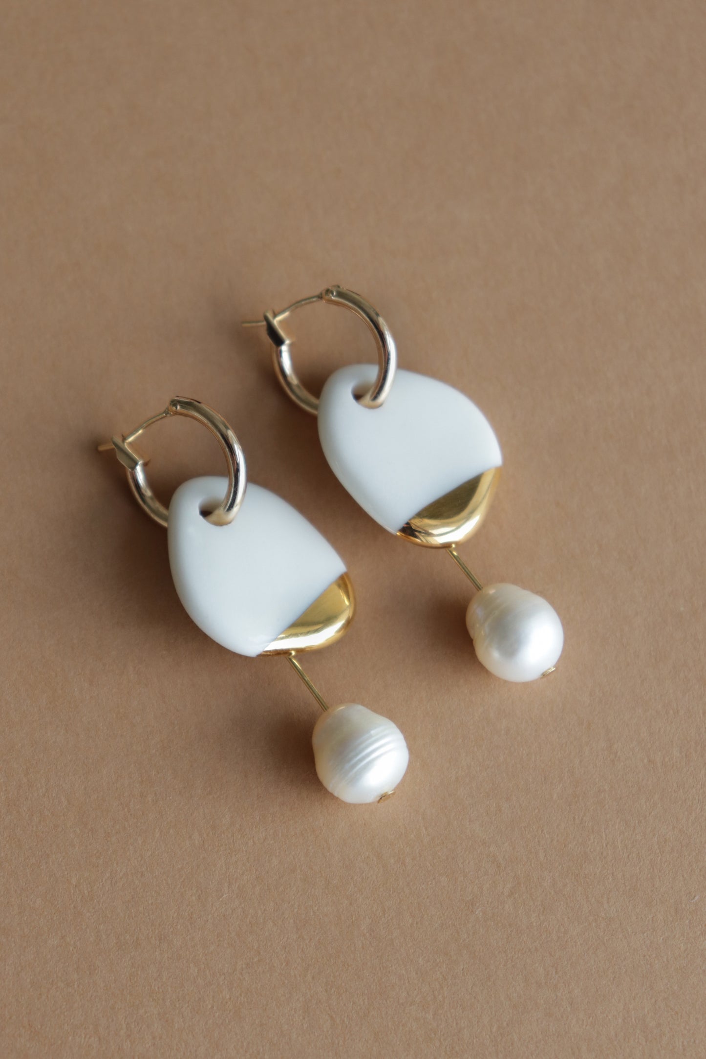 The Arp Pearl Hoops by Rozenthal Jewelry feature gold hoops with white geometric accents and dangling round freshwater pearls, beautifully displayed on a brown background—a striking handmade accessory.