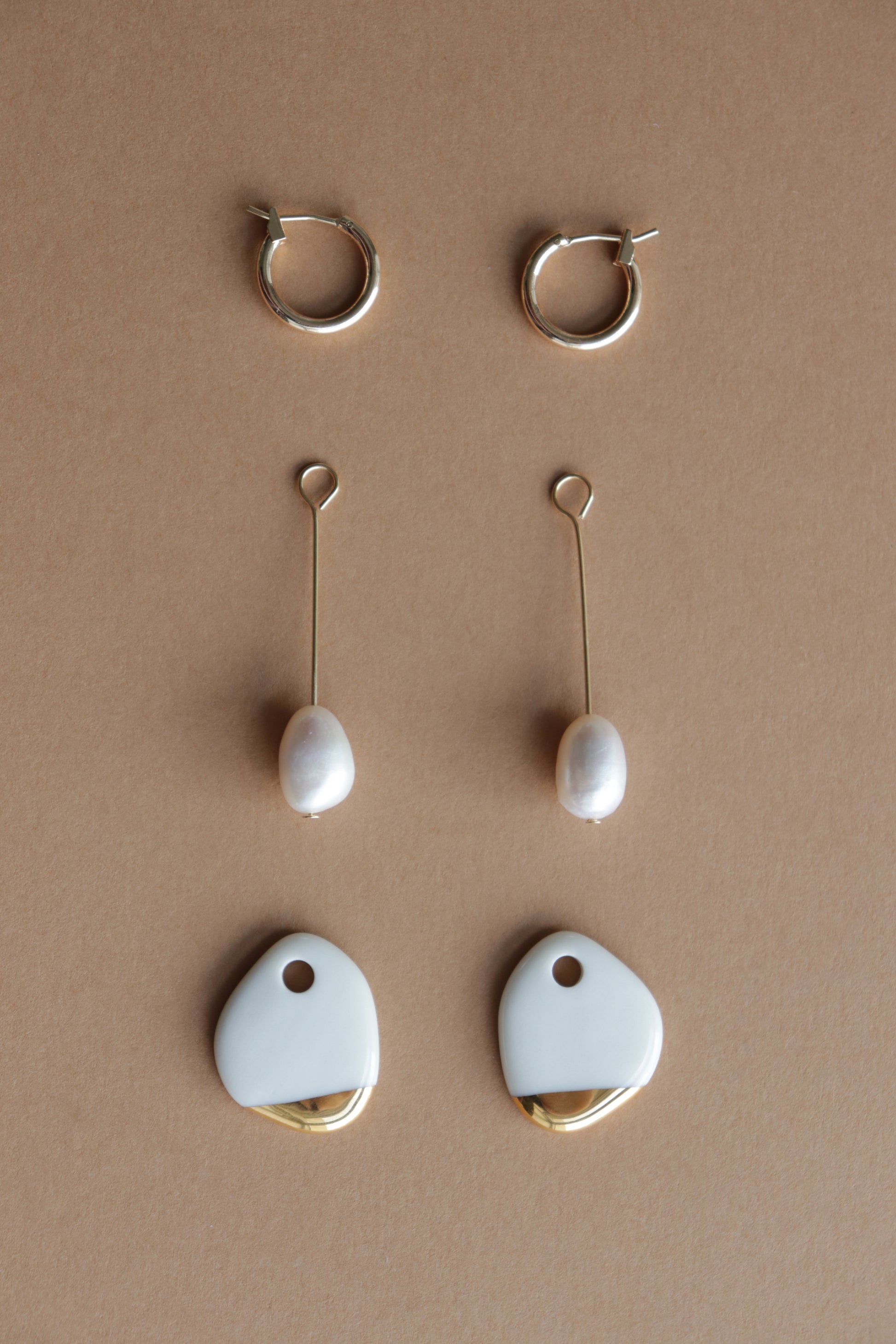 Three pairs of earrings from Rozenthal Jewelry: gold hoops (Arp Pearl Hoops) at the top, freshwater pearl earrings in the middle, and abstract gold and white statement pieces at the bottom on a beige background.