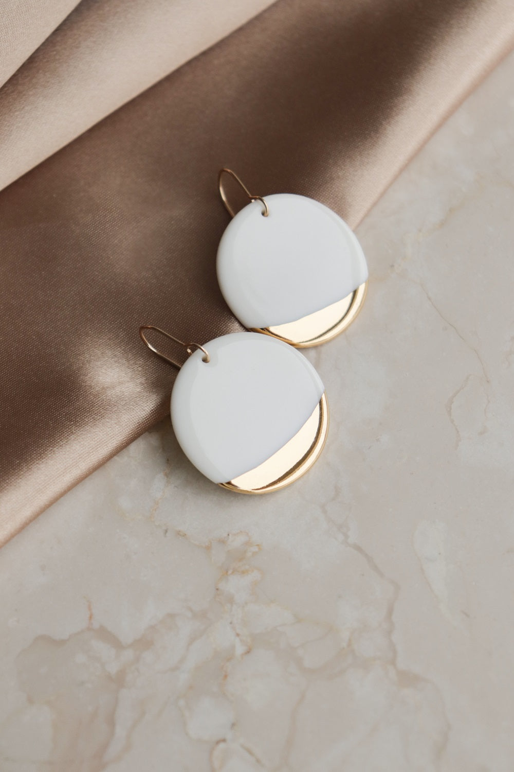 Round Earrings by Rozenthal Jewelry feature modern white and gold geometric shapes in a minimalist design, shown on marble beside soft tan fabric.
