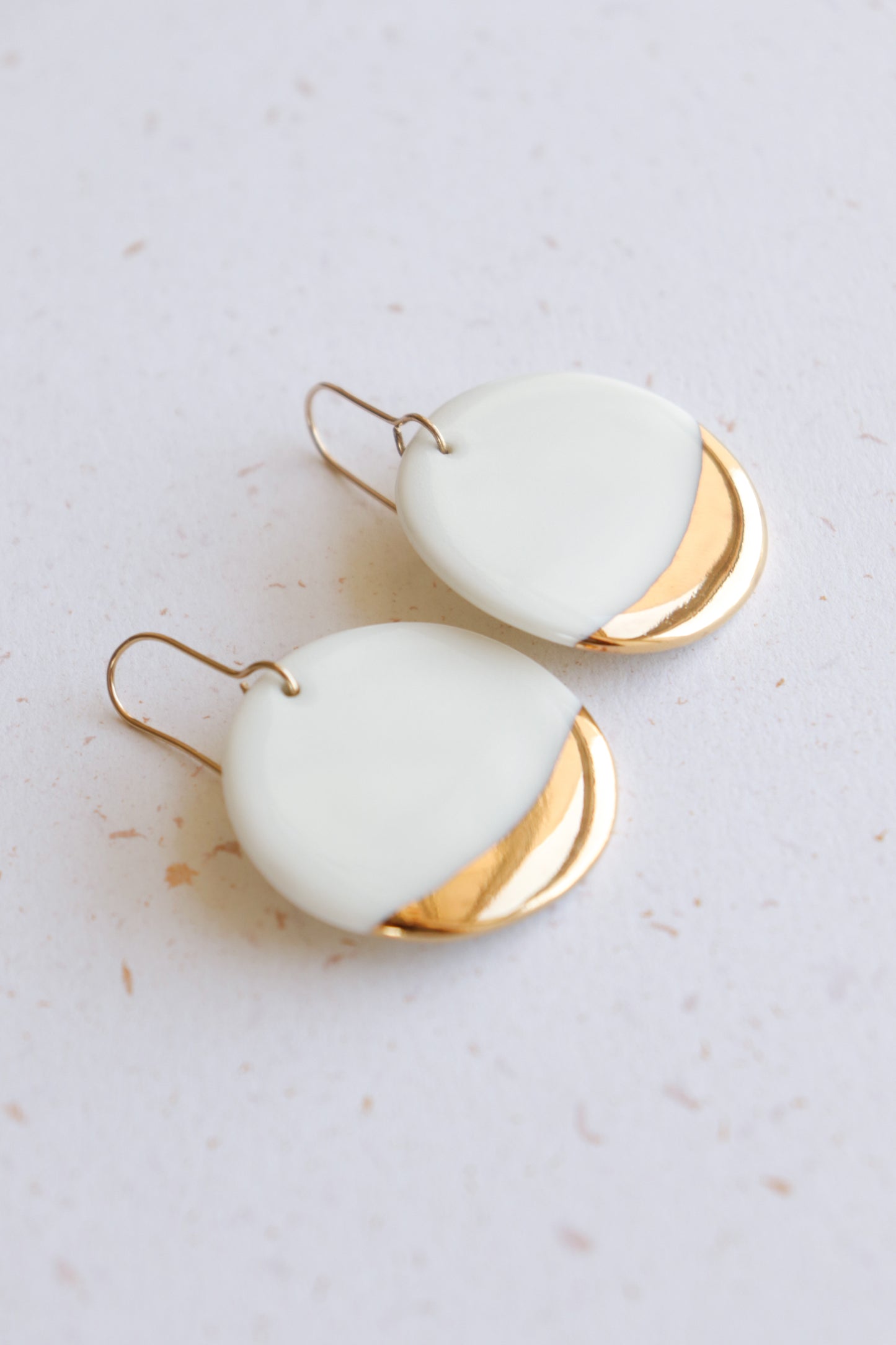 Round Earrings by Rozenthal Jewelry feature minimalist white and gold halves with a modern, smooth design and gold hooks. Shown lying on a light speckled surface, these dangle earrings add a stylish touch.