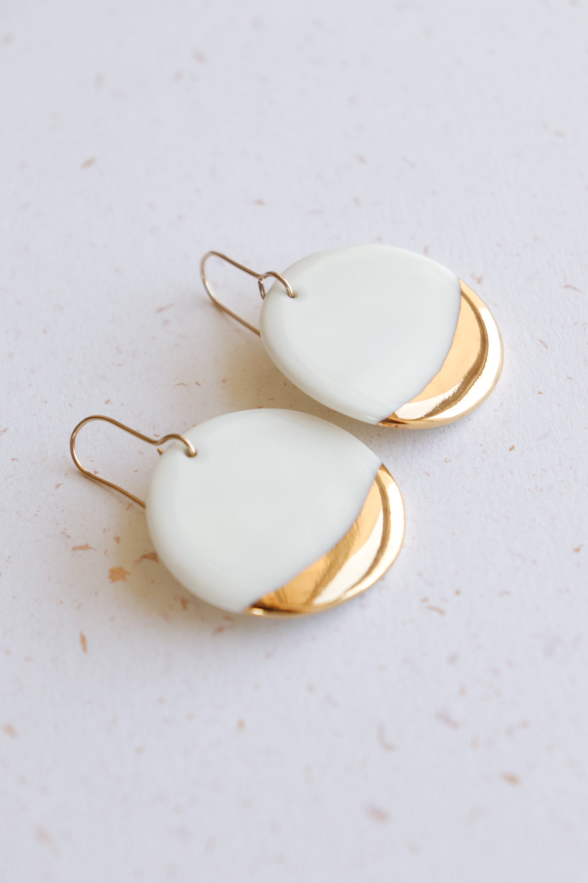 Round Earrings by Rozenthal Jewelry feature minimalist white and gold halves with a modern, smooth design and gold hooks. Shown lying on a light speckled surface, these dangle earrings add a stylish touch.