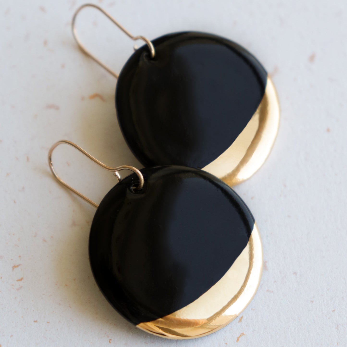 Rozenthal Jewelrys Round Earrings feature a glossy black upper half and shiny gold lower half, creating a minimalist look. Shown on a light, speckled surface, these dangle earrings offer modern elegance.